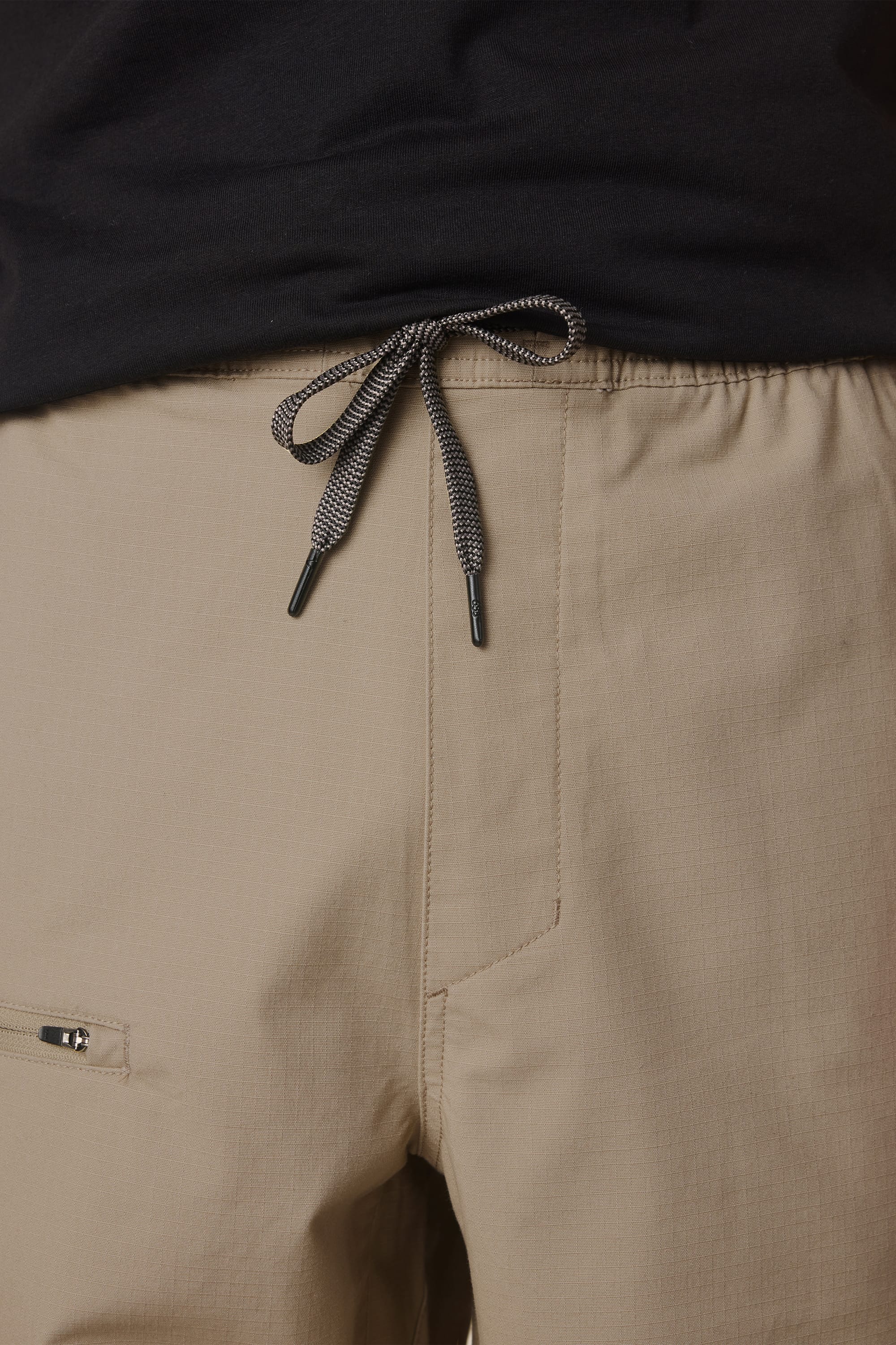 Close-up of a person wearing the 686 Men's Free Loader Pant - Loose Fit in beige, featuring a visible knotted drawstring and waistband, along with part of a zippered pocket on the nylon woven pant. The pants are paired with a black shirt.