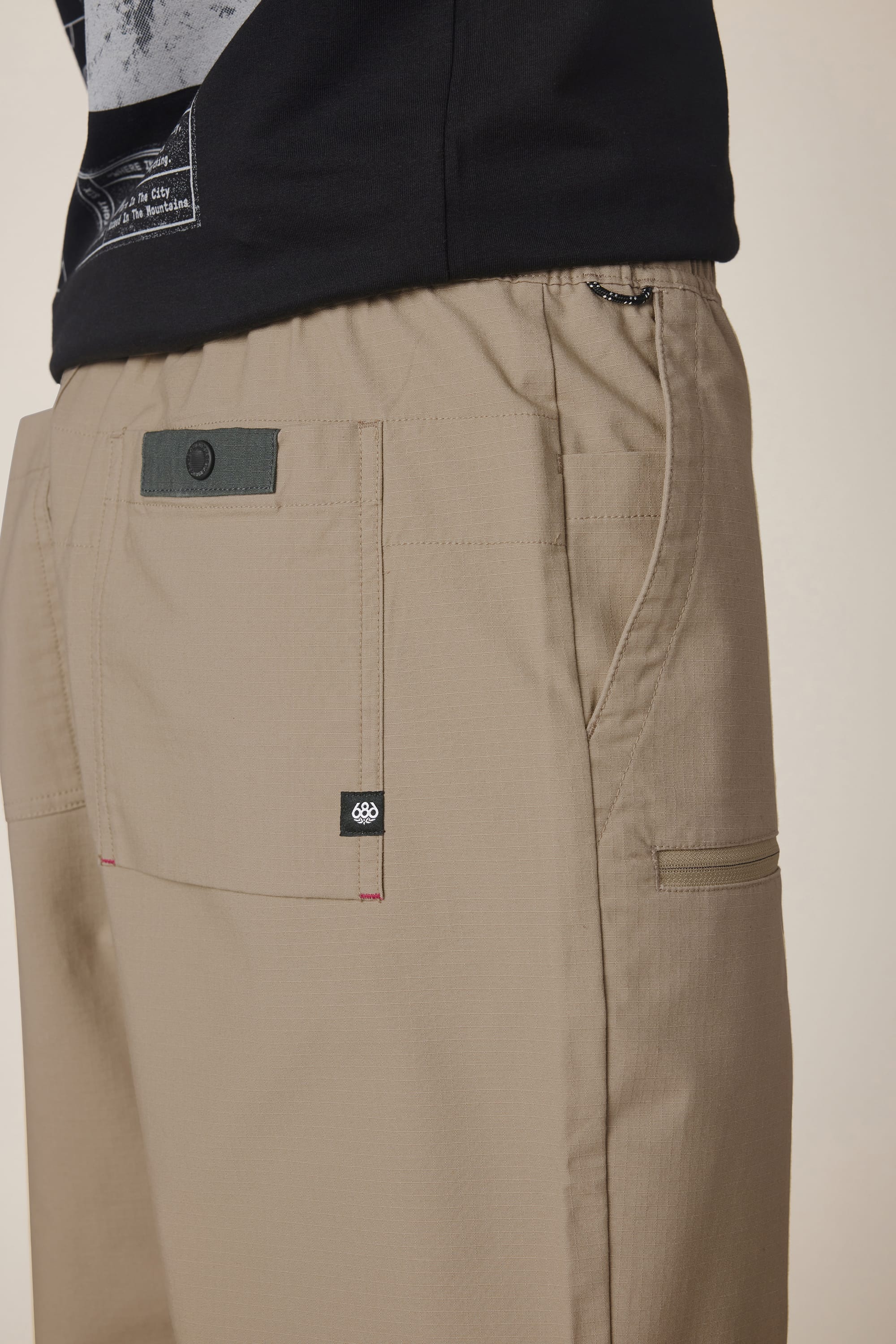A close-up of a person wearing the 686 Men's Free Loader Pant - Loose Fit in beige, featuring a back pocket with a buttoned flap and small black brand label, paired with a black shirt. These pants by 686 are crafted from durable nylon woven fabric for long-lasting wear.