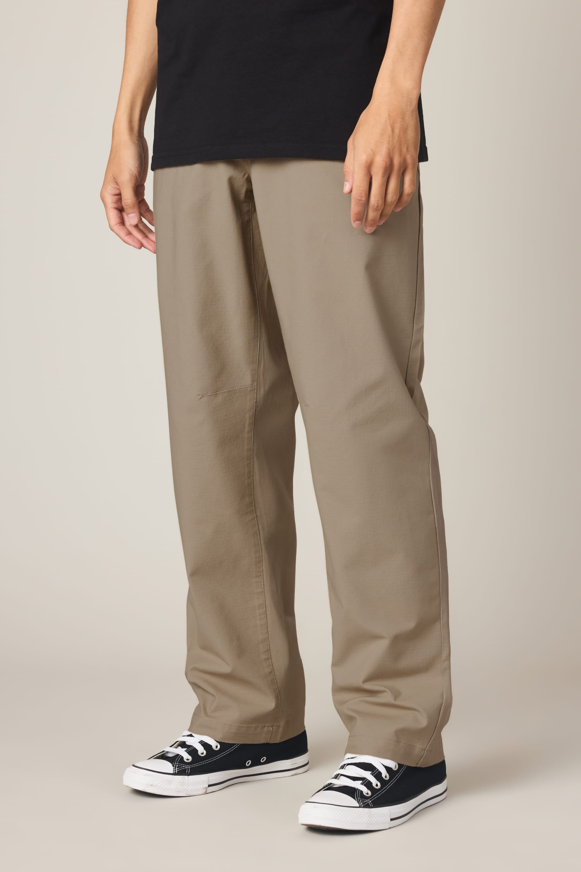 A person wears the 686 Men's Free Loader Pant - Loose Fit in beige, paired with a black shirt and black low-top sneakers with white soles, standing against a plain, light background.