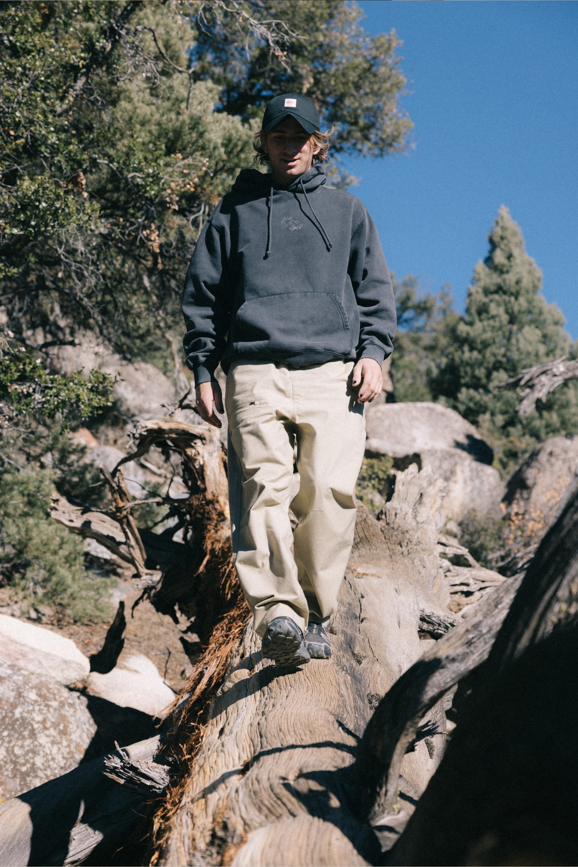A person wearing a black hoodie, a 686 Men's Free Loader Pant - Loose Fit by 686, and a cap walks on a large fallen tree trunk in a rocky, forested area under a clear blue sky.