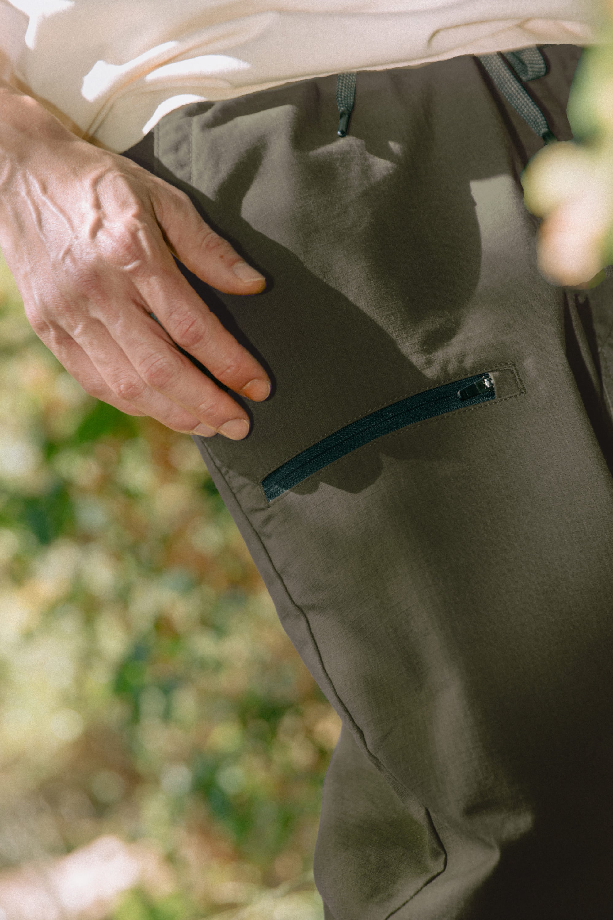 A close-up of a person’s hand resting near the front pocket zipper of the 686 Men's Free Loader Pant - Loose Fit in olive green, with sunlight and blurred greenery in the background.