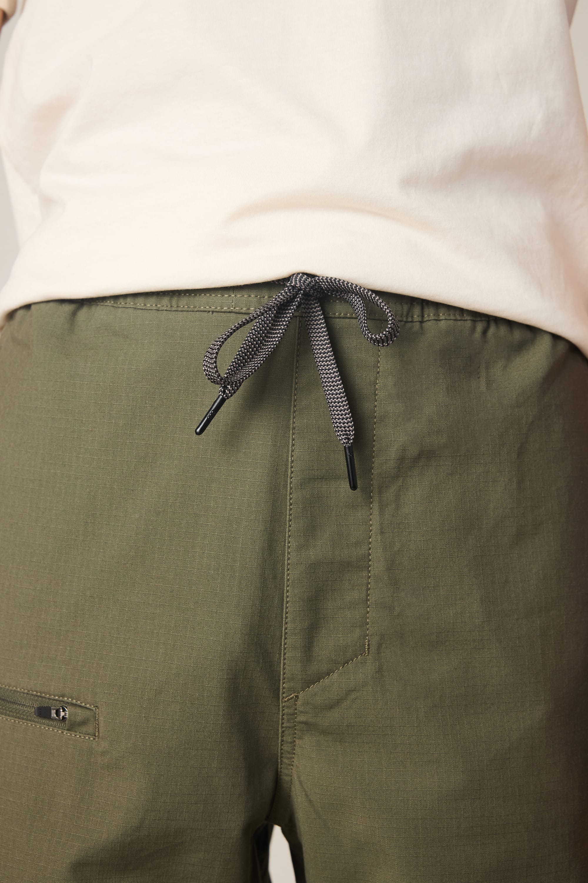 Close-up of a person wearing 686 Men's Free Loader Pant - Loose Fit in olive green with a gray and black drawstring, paired with a white shirt. The image highlights the nylon woven fabric at the waist and upper legs.