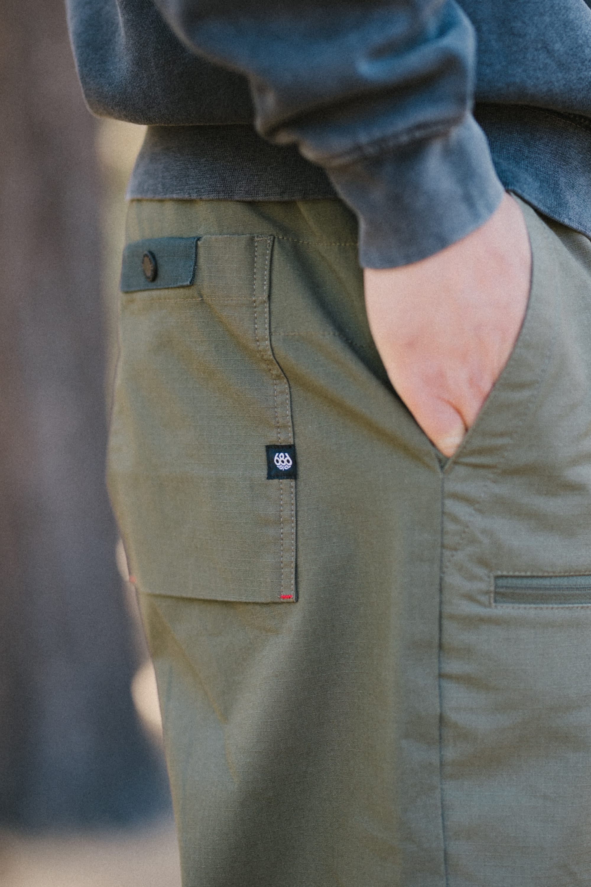 A person is seen wearing the 686 Men's Free Loader Pant - Loose Fit in olive green, featuring a flap pocket with a dark button and a durable nylon woven finish. They stand with one hand in the pocket, paired with a dark sweatshirt against a blurred background.