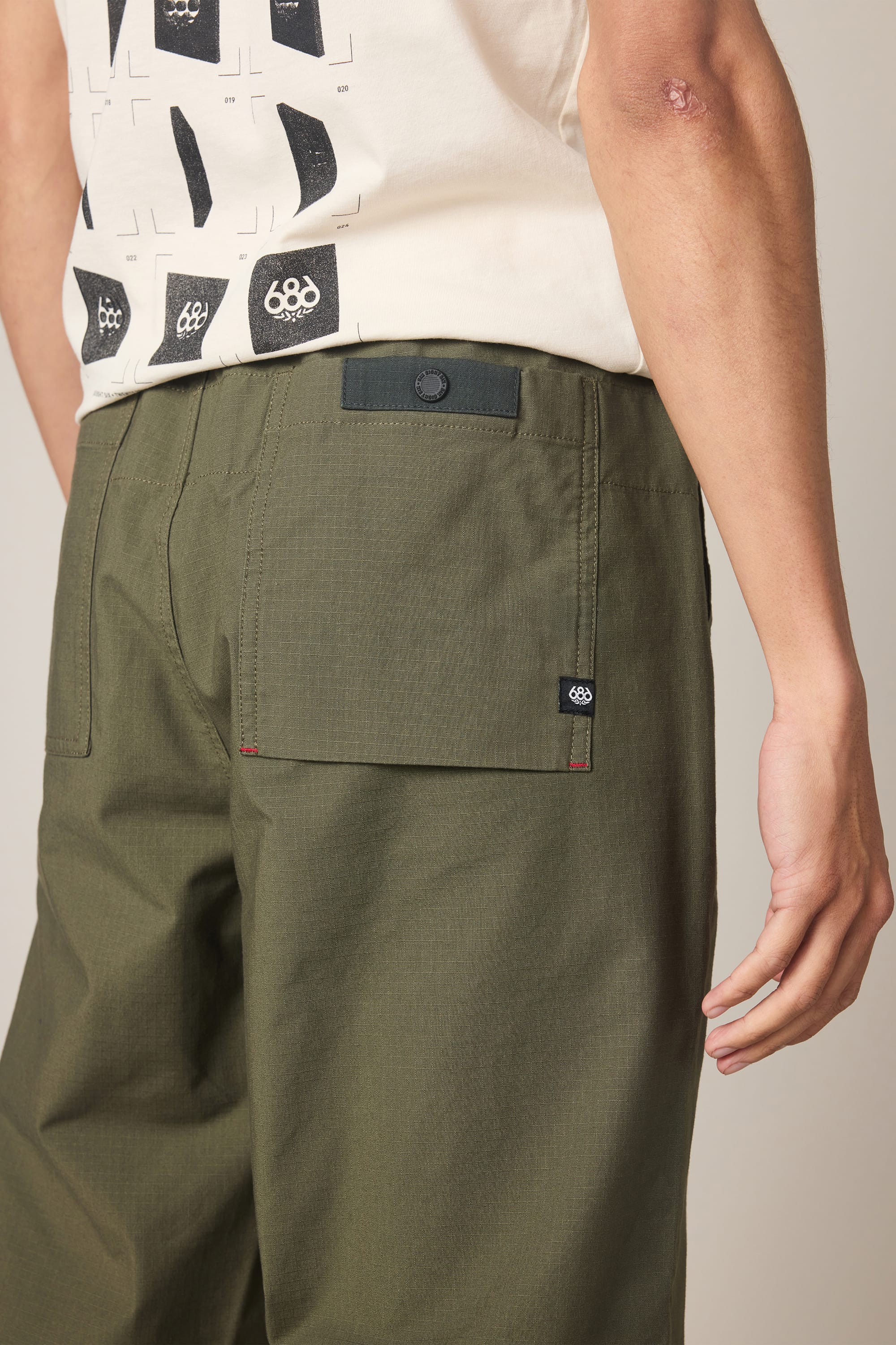 A person wears the 686 Men's Free Loader Pant - Loose Fit in olive green, featuring back pockets, paired with a white graphic t-shirt. The photo highlights the back of the nylon woven pant from 686 and shows part of the person’s right arm.