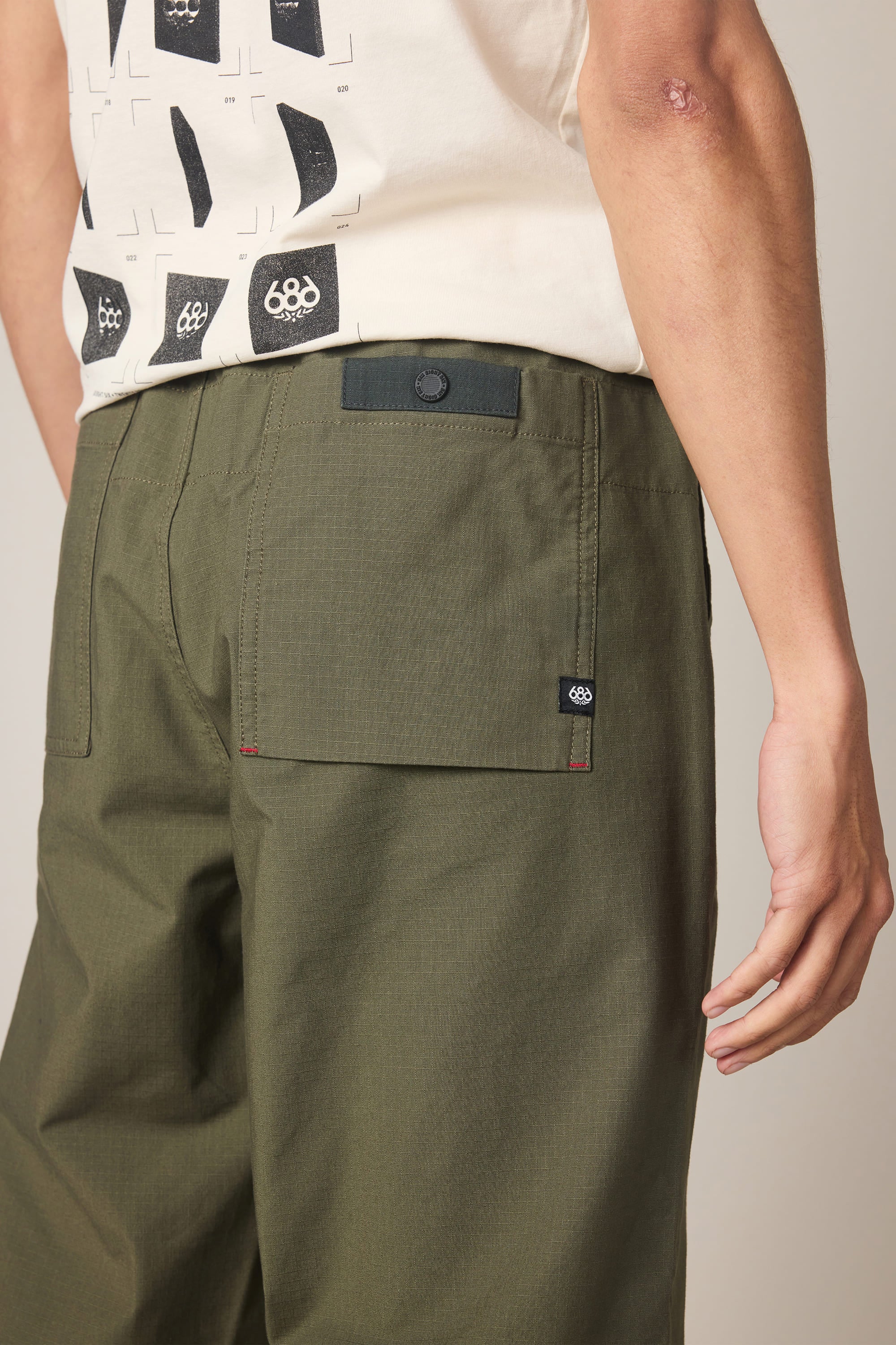 A person wearing the 686 Men's Free Loader Pant - Loose Fit in olive green, featuring back pockets, paired with a cream shirt with black graphic designs; their right arm is visible with a scar or mark near the elbow.