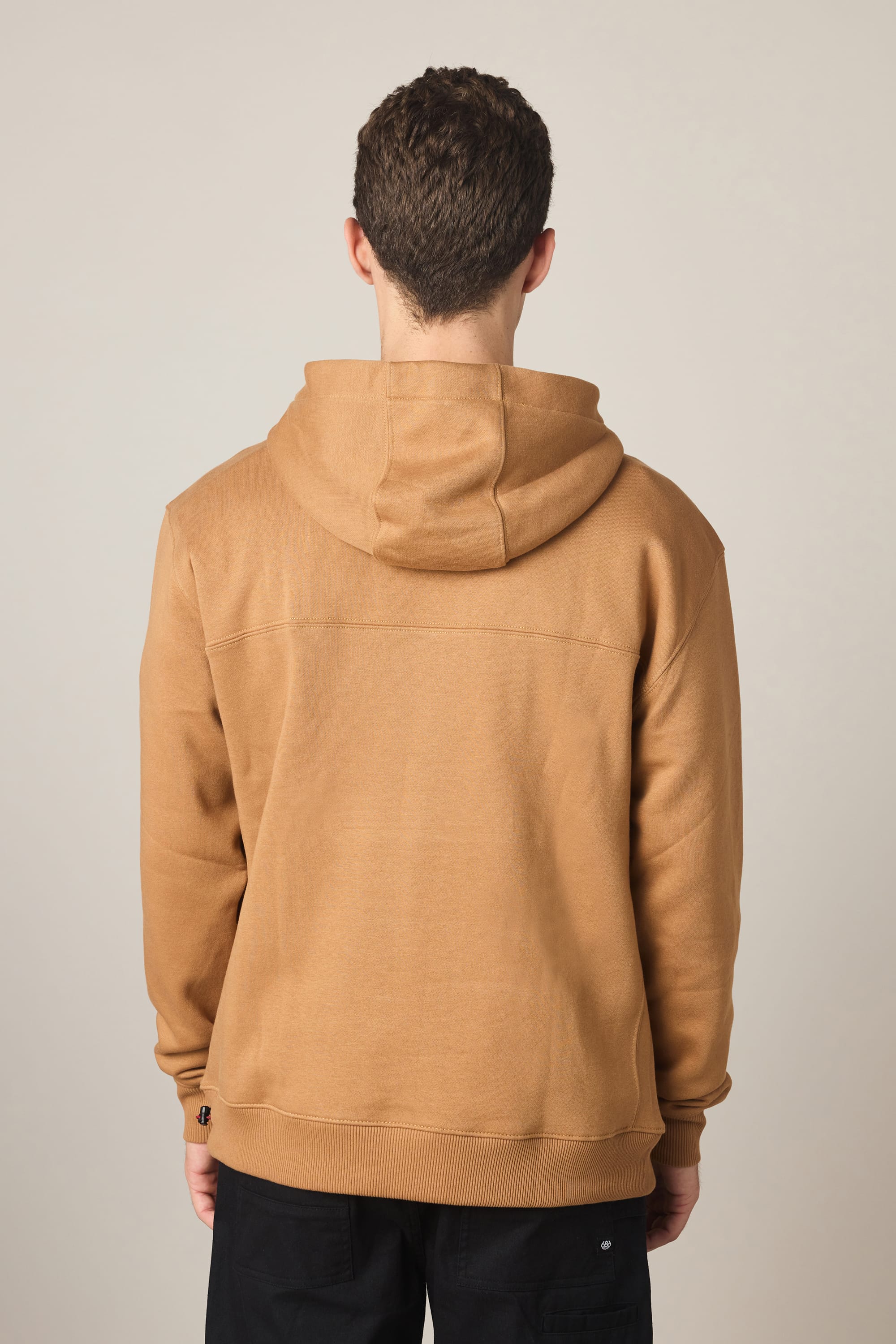A person with short curly hair is shown from the back, wearing the 686 Men's All Season Hoody in tan and black pants, standing against a plain light-colored background.