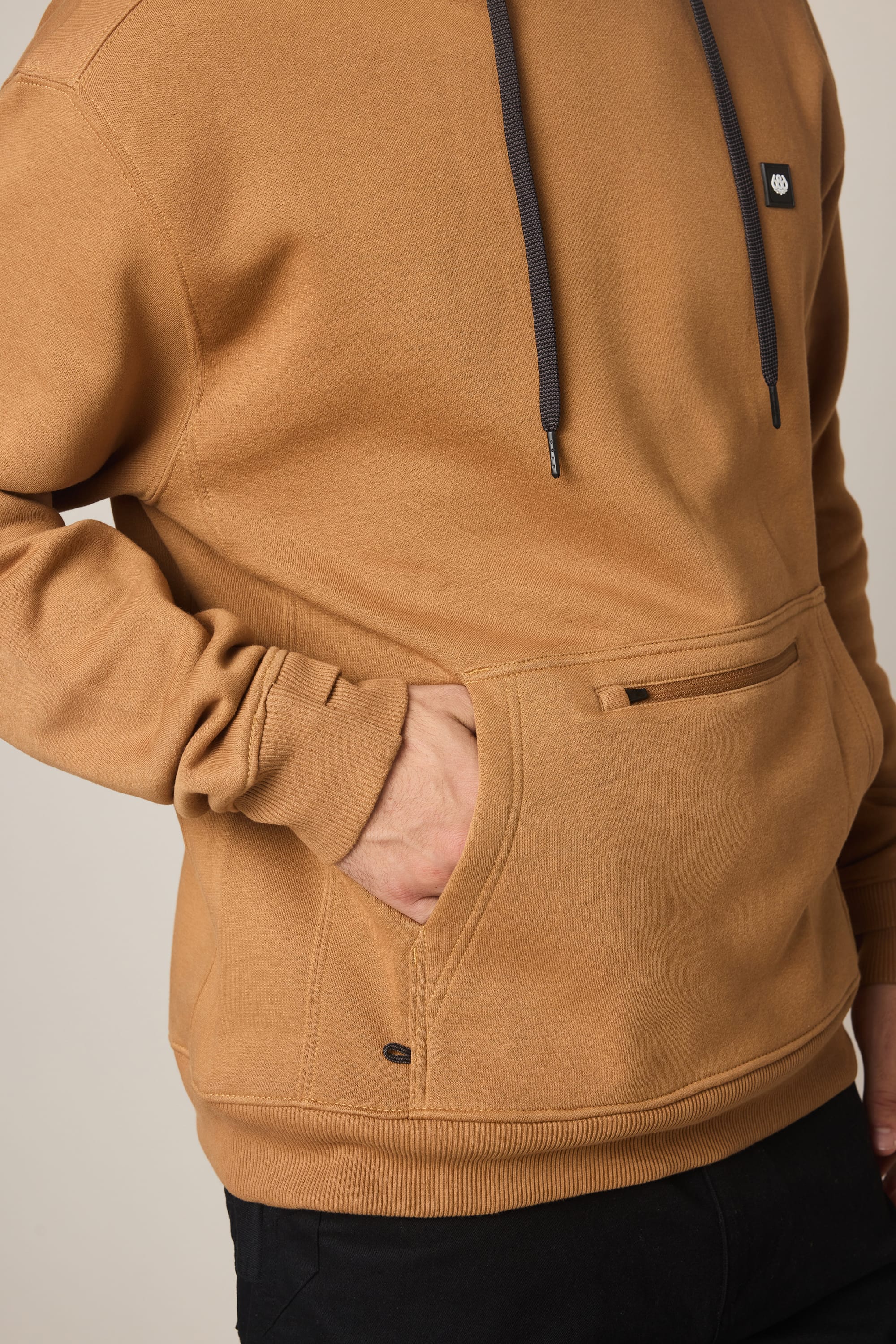 A person is shown from the torso up, wearing the 686 Men's All Season Hoody in brown. One hand rests in the front pocket of the cotton sweatshirt. The hoodie from 686 features dark drawstrings and a small black patch on the chest, with only part of one arm visible.