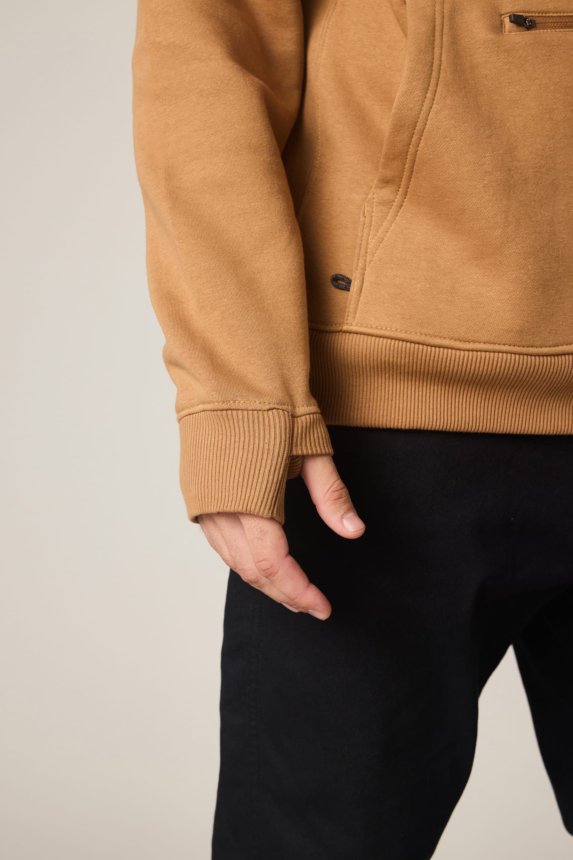 A person is shown from the shoulders down wearing the 686 Men's All Season Hoody in tan, featuring ribbed cuffs, with black pants and their left hand relaxed at their side.
