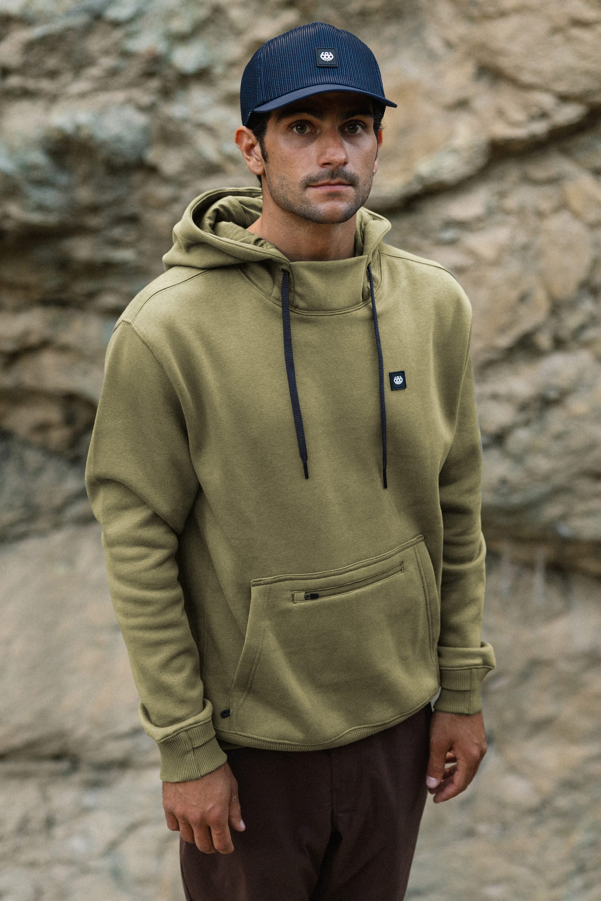 A man in a 686 Men's All Season Hoody and dark cap stands outdoors in front of a rocky background, looking at the camera with a neutral expression.