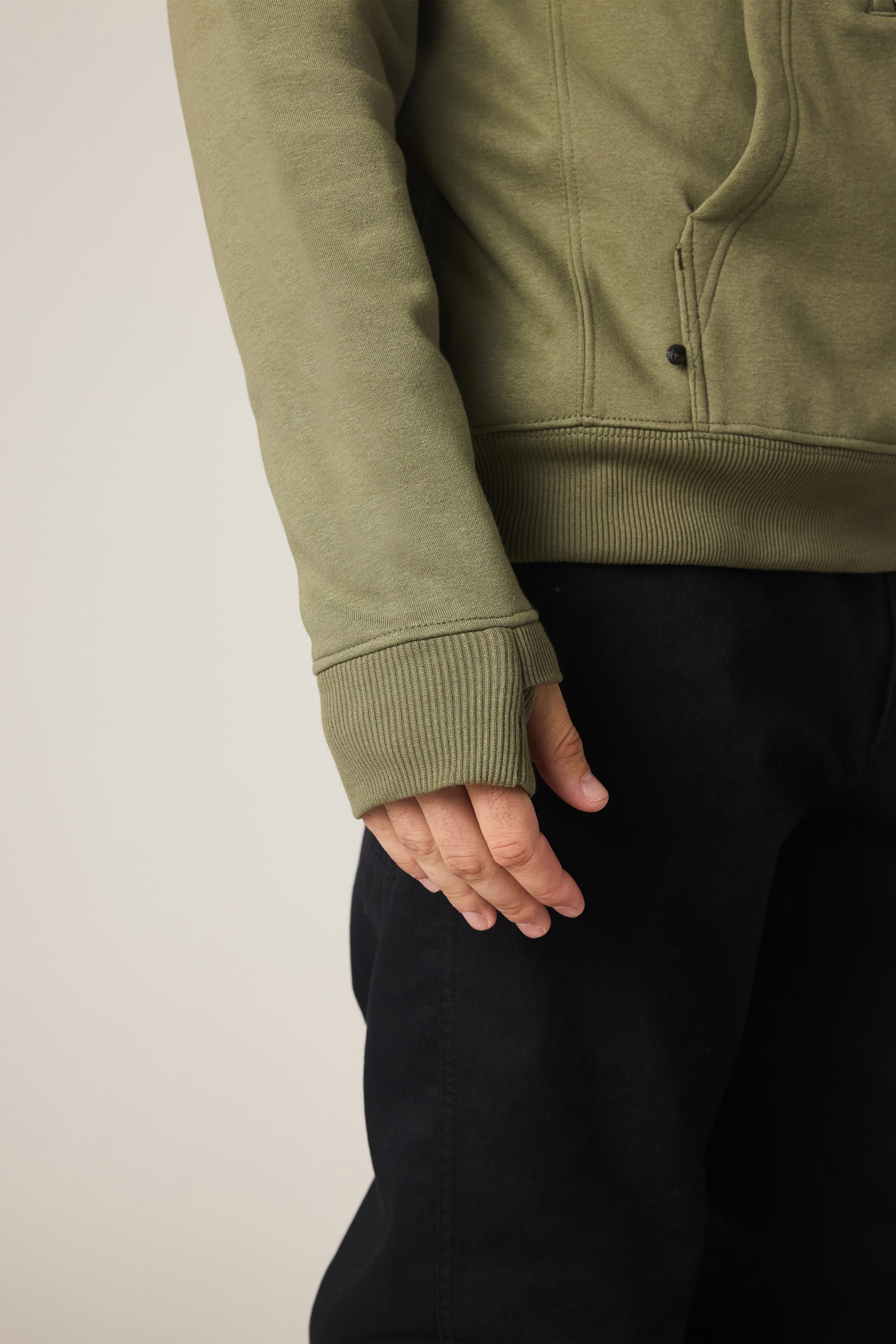 A person shown from the shoulders down wears a 686 Men's All Season Hoody in light olive green with ribbed cuffs and black pants, left arm relaxed at their side against a plain background.