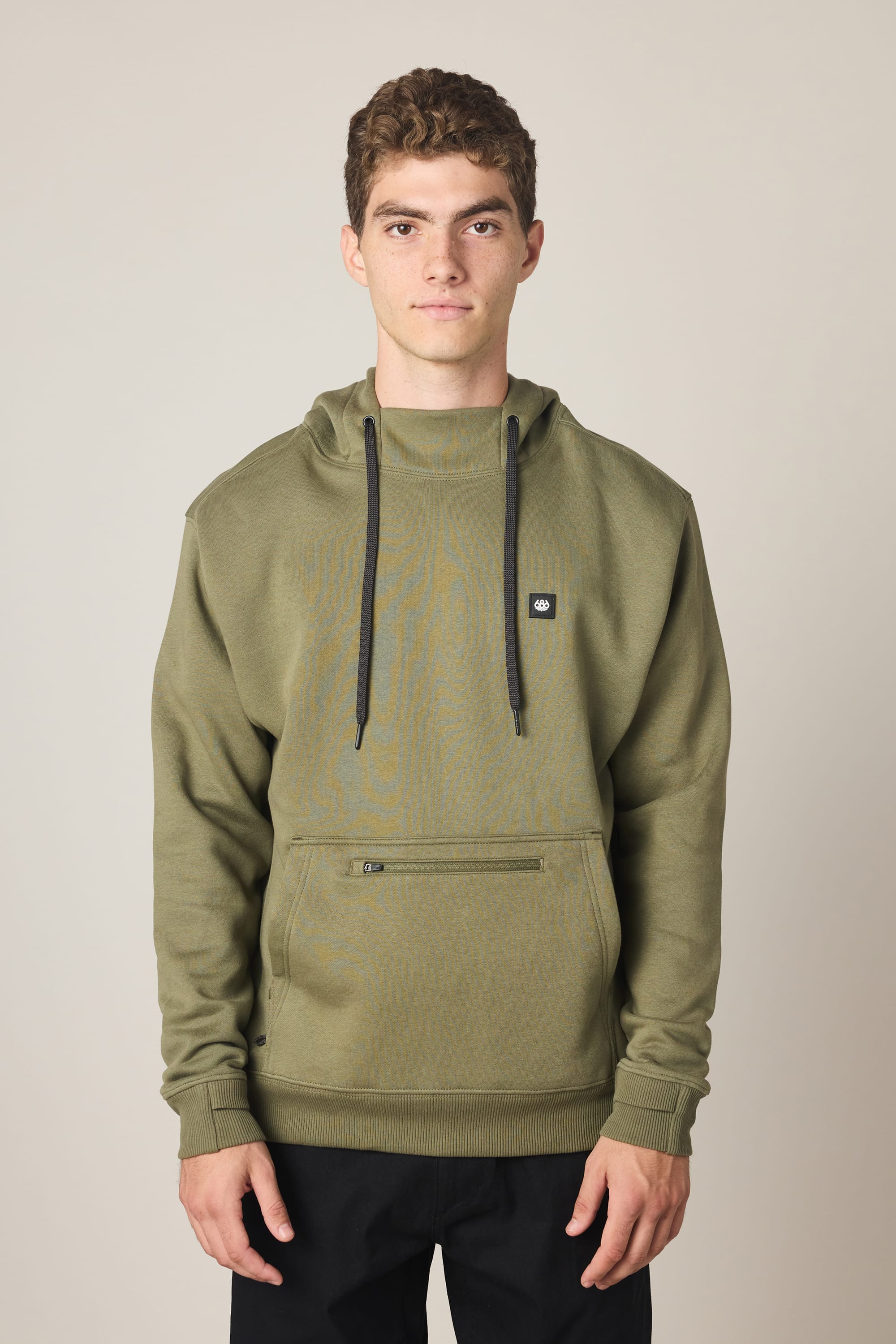 A young man with short curly hair models the 686 Men's All Season Hoody in olive green, featuring a front pocket and black drawstrings. He stands against a plain light background with a neutral expression and arms by his sides.