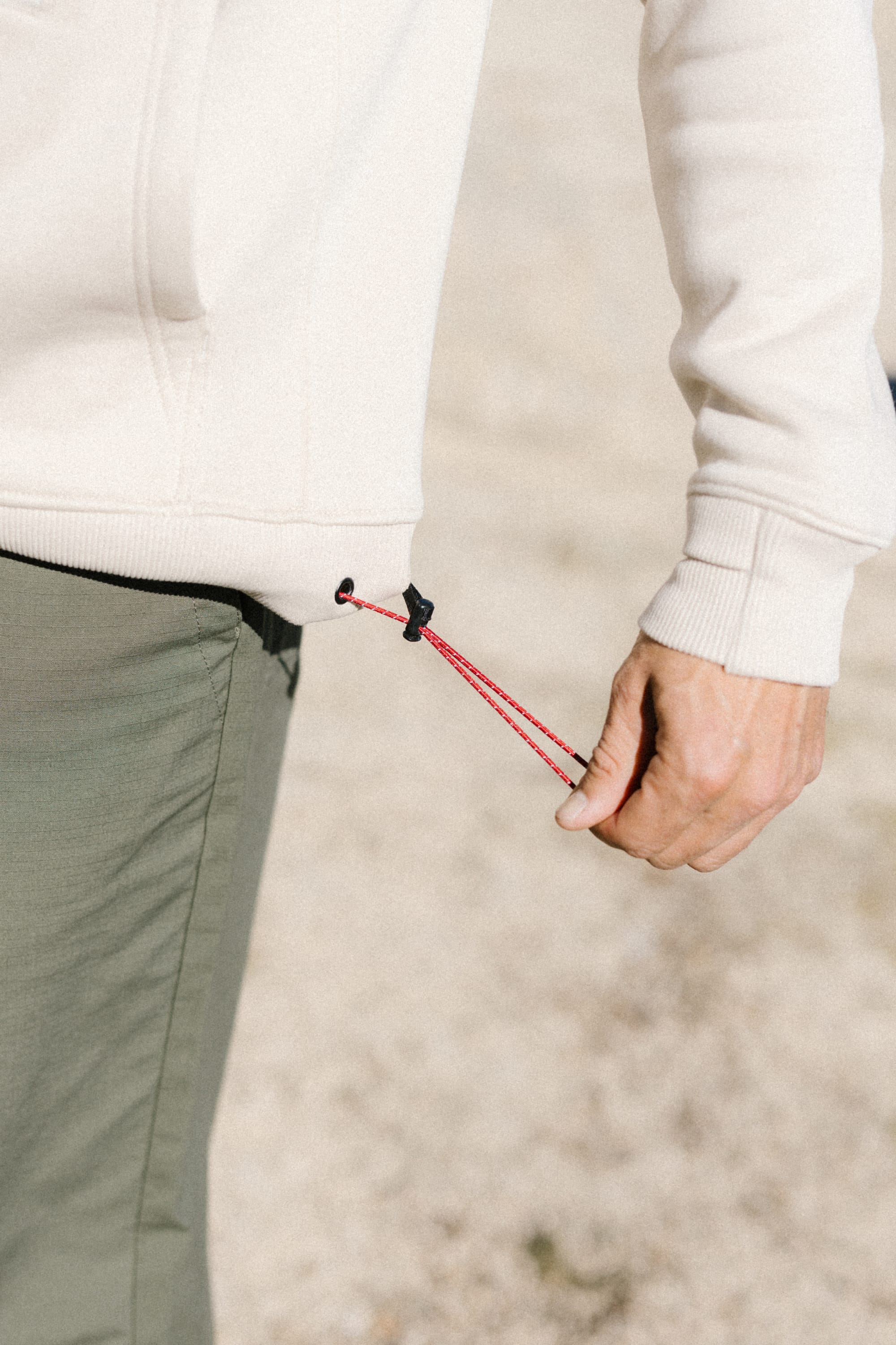 A person in green pants and the 686 Men's All Season Hoody in cream cotton tightens a red drawstring at the hem of the sweatshirt.