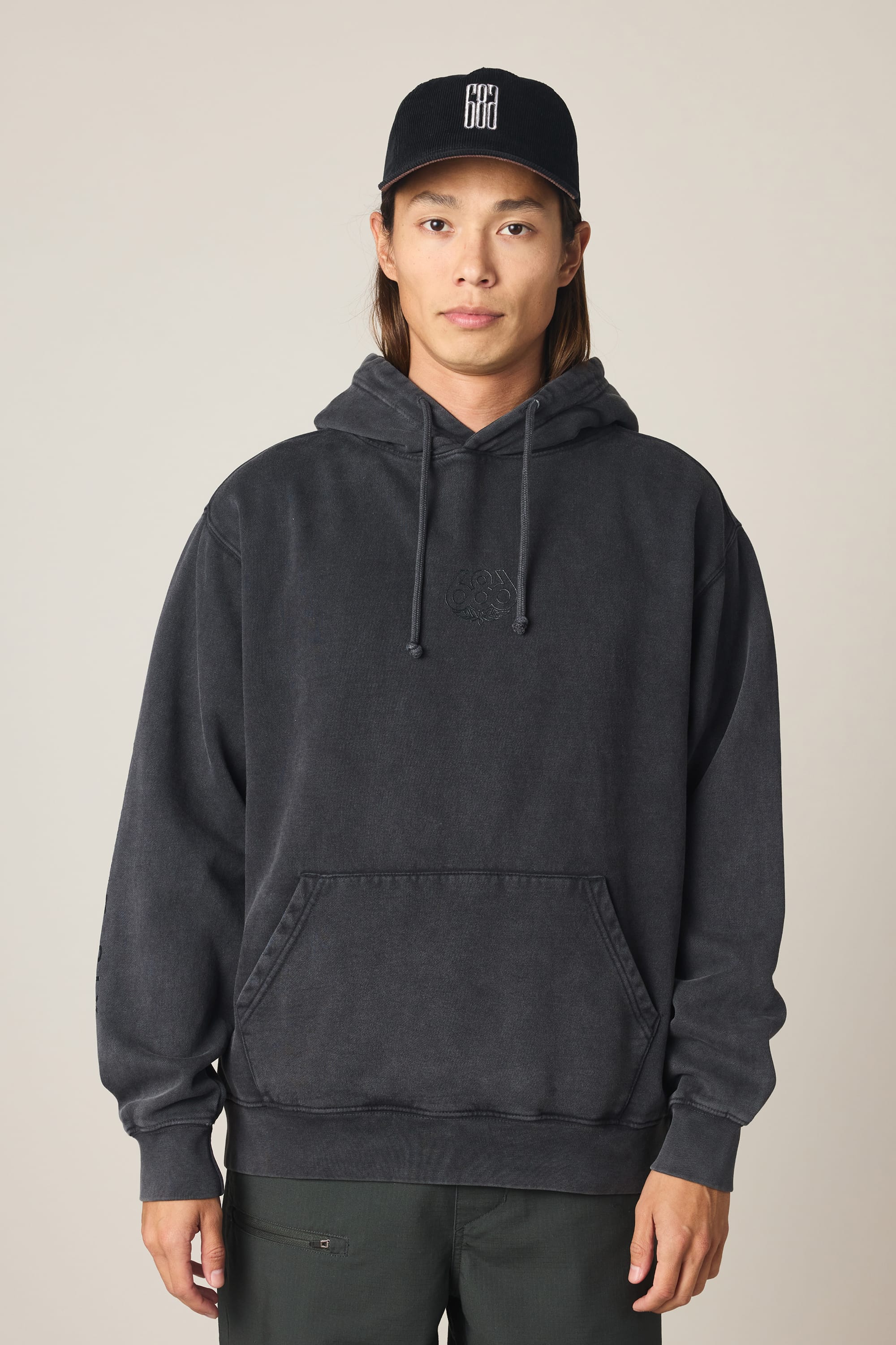 A person with long hair wears a 686 Men's High Def Relaxed LS Hoody in black and a black cap with white lettering, standing against a plain light background.