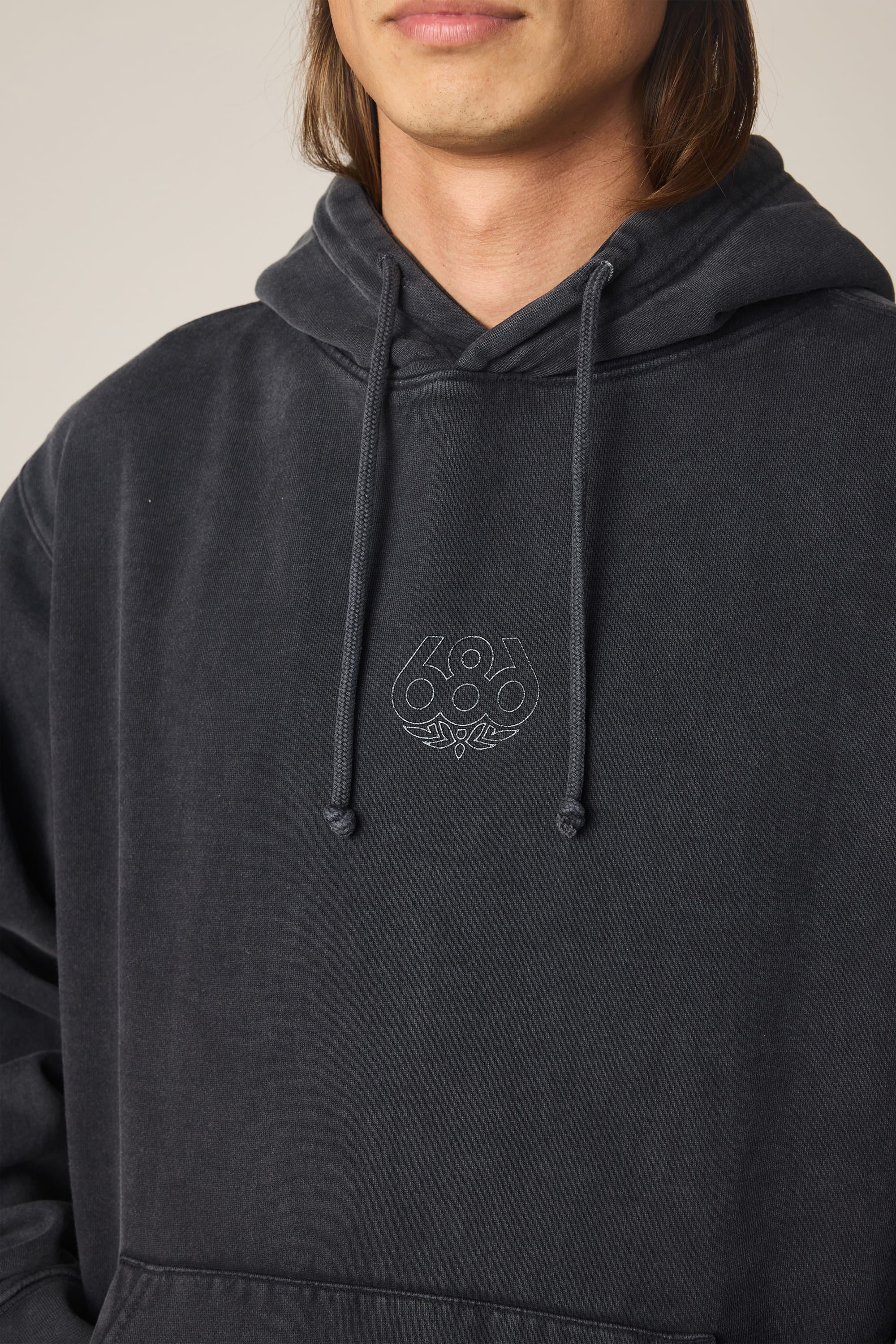 A person is wearing the 686 Men's High Def Relaxed LS Hoody, a black cotton blend sweatshirt from 686 featuring a small embroidered logo on the chest. Their face is partially visible against a plain, neutral background.