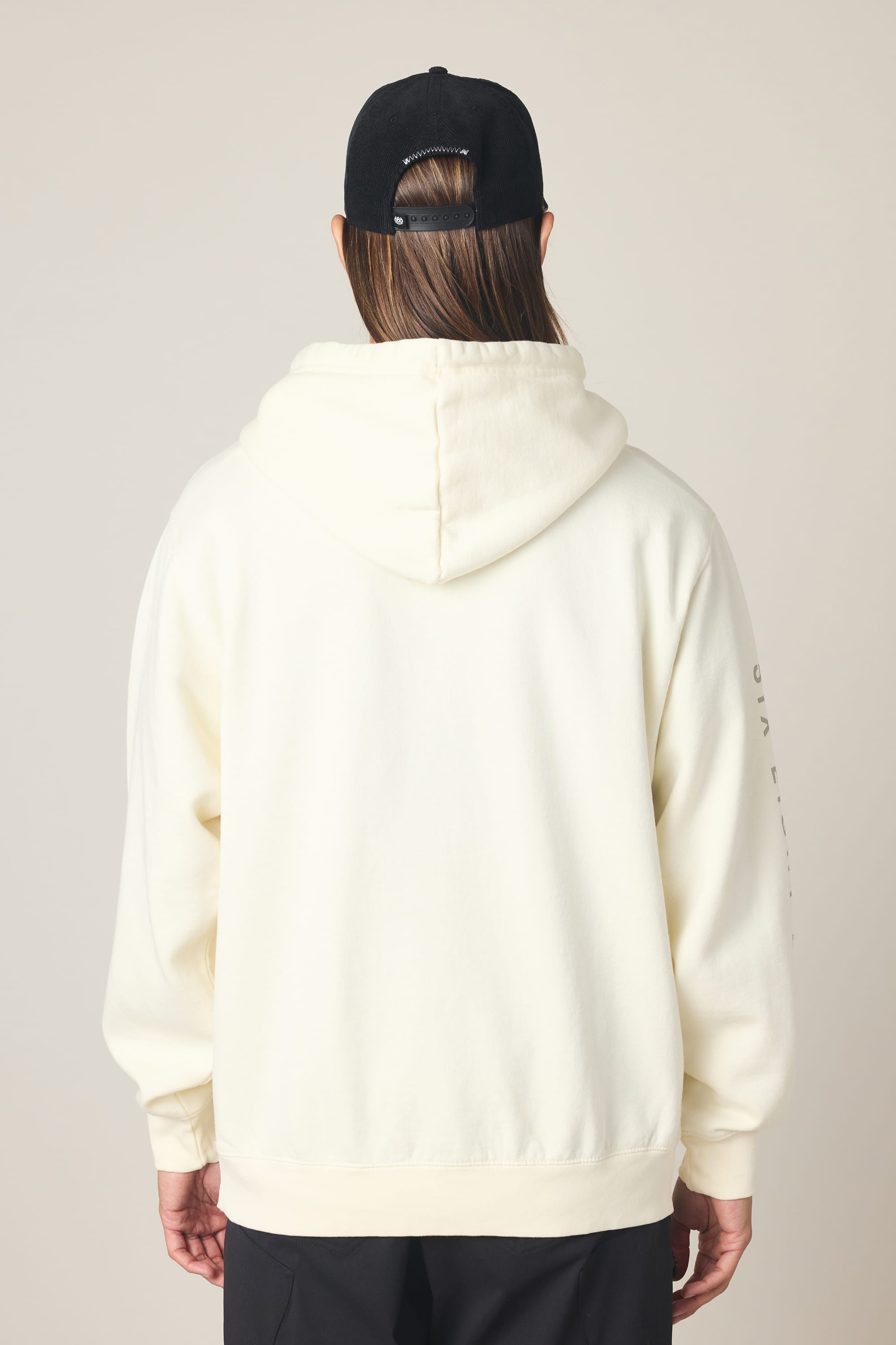 A person with long hair, wearing a black baseball cap, 686 Men's High Def Relaxed LS Hoody by 686, and black pants stands with their back to the camera against a plain background.
