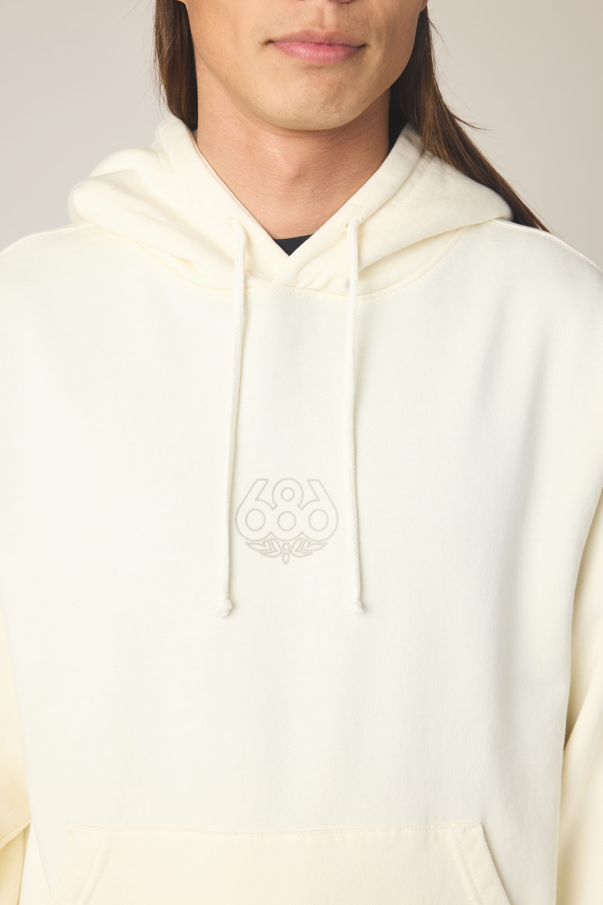 A person wearing the 686 Men's High Def Relaxed LS Hoody, an off-white cotton blend hoodie featuring a subtle embroidered 686 logo and a small design. The photo shows the upper torso, part of the face, and long brown hair.