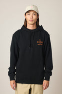 A person stands facing forward against a neutral background, wearing the 686 Men's Unwork Spec Athletic LS Hoody in black with orange graphics by 686, paired with a light-colored baseball cap and beige pants.