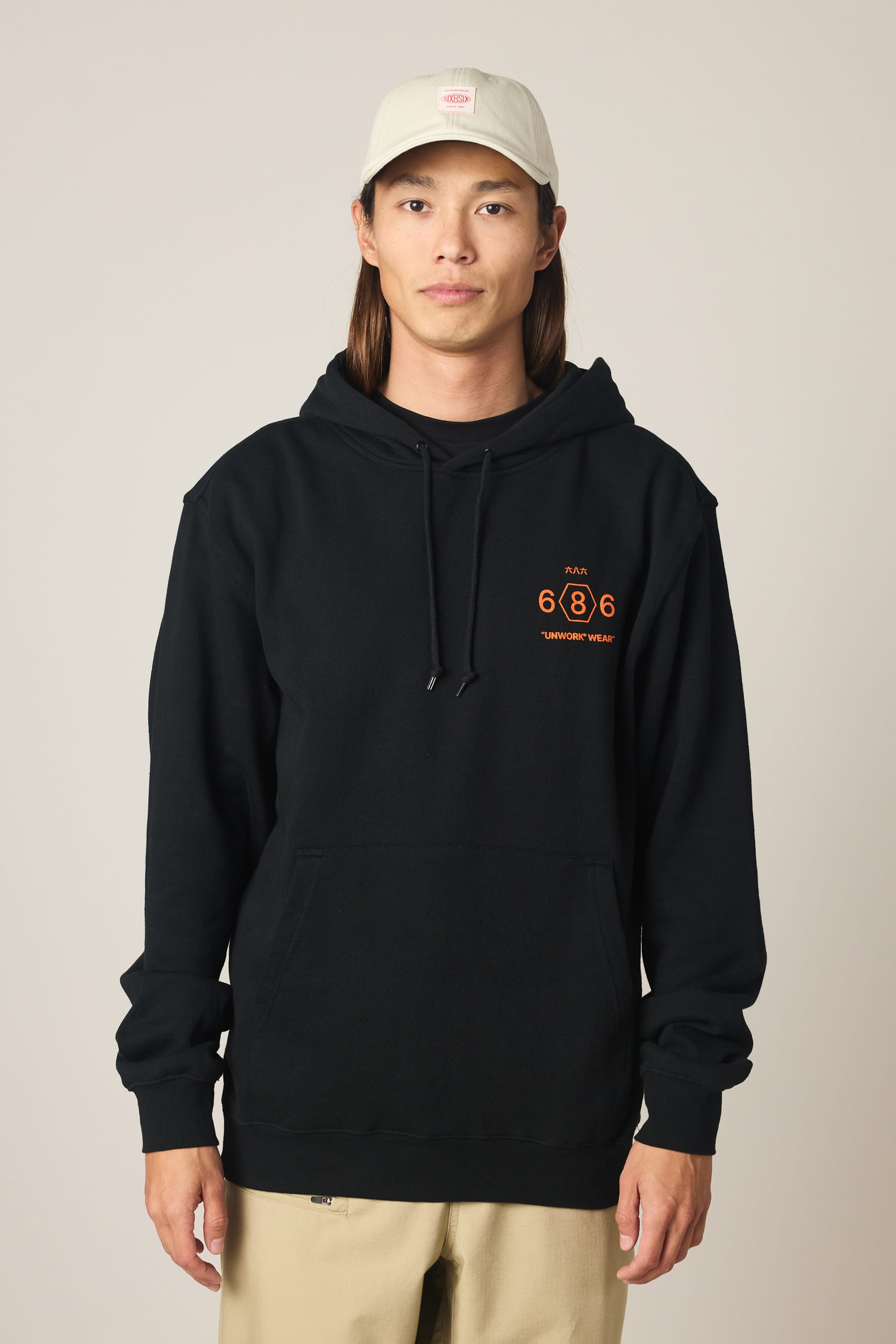A person wearing the 686 Men's Unwork Spec Athletic LS Hoody in black with orange text, tan pants, and a light beige baseball cap stands facing forward against a plain light background.