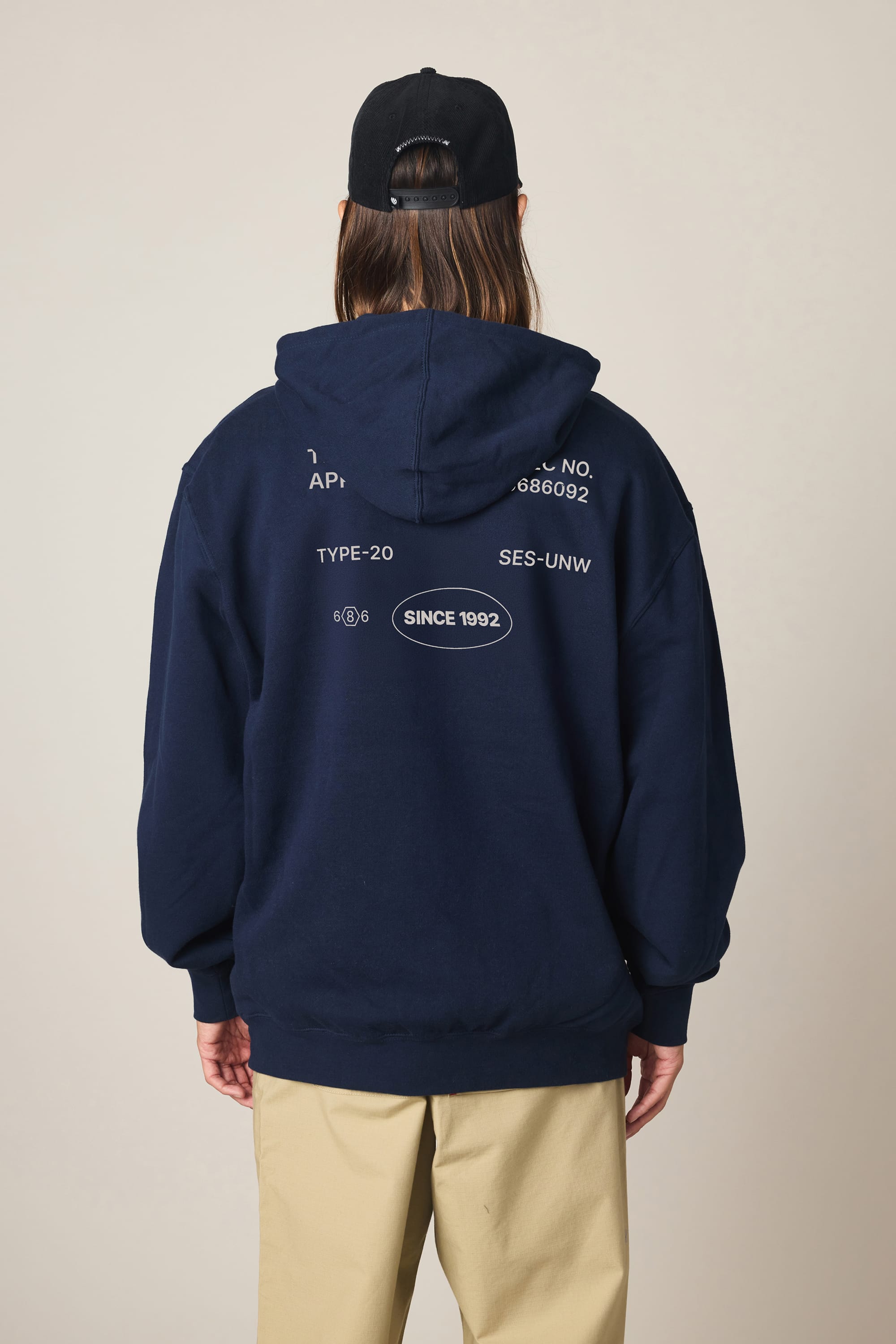 Model wears the 686 Men's Unwork Spec Athletic LS Hoody in navy with white graphics on the back, paired with beige pants and a black cap, standing against a plain light background, facing away from the camera.