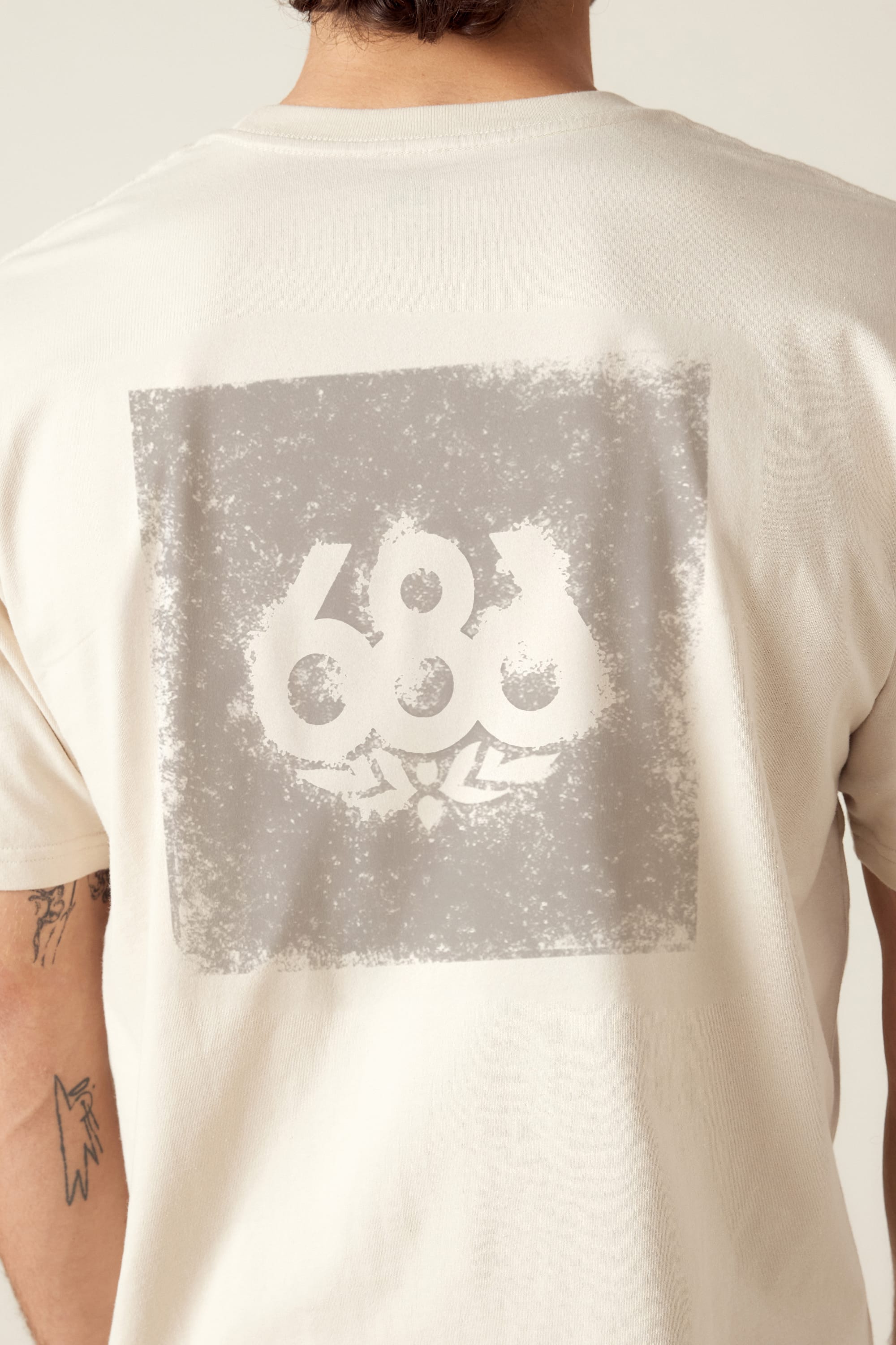 A person wears the 686 Knockout Imprint Authentic S/S Tee, a light-colored mens t-shirt with a faded square 686 graphic on the upper back. Only their upper back and arms are visible. Made by 686 from 100% cotton for comfort.