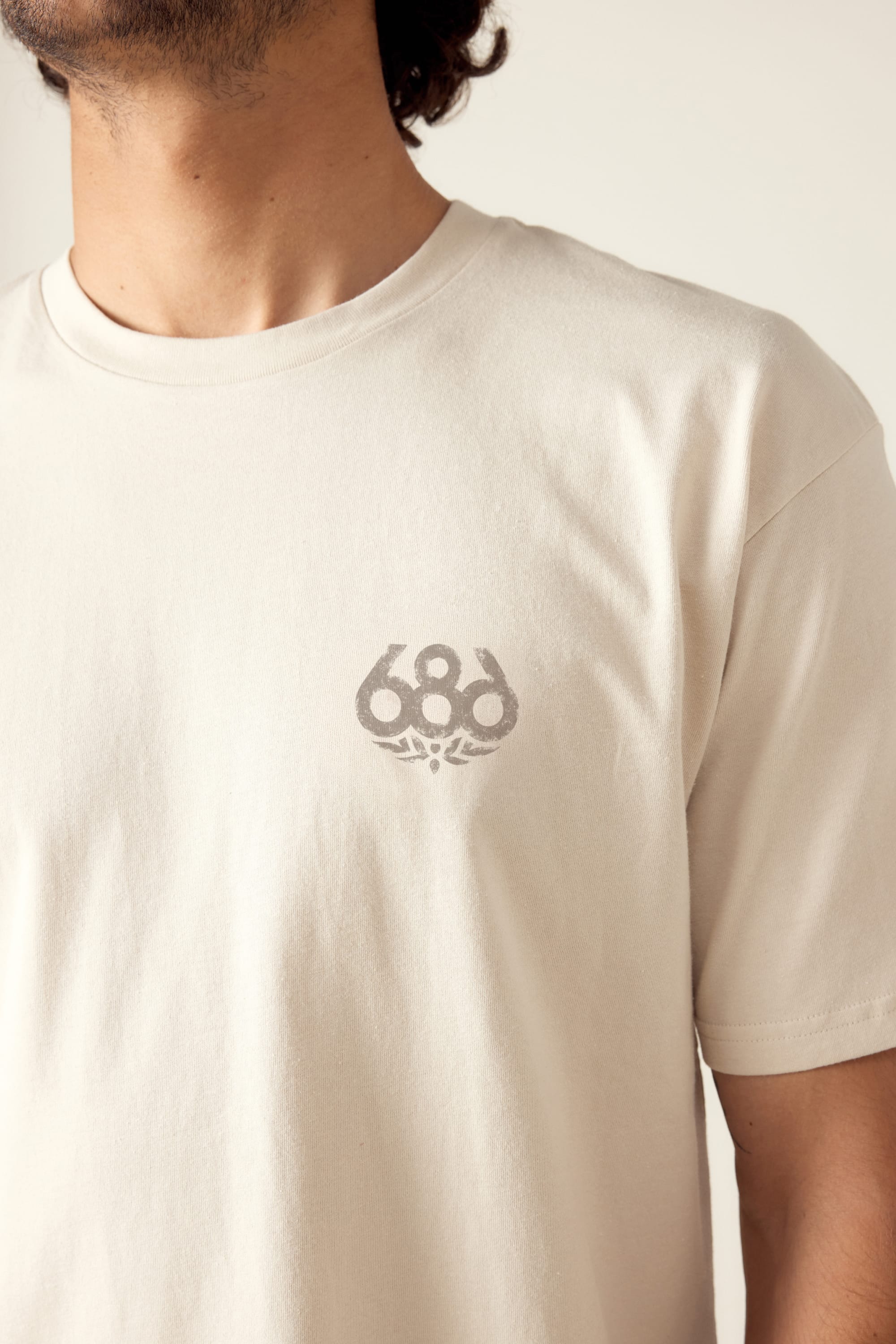 A person wears the 686 Knockout Imprint Authentic S/S Tee, a light beige mens cotton t-shirt featuring a small gray 686 logo on the left chest. The image captures their upper torso, part of their face, and an arm.