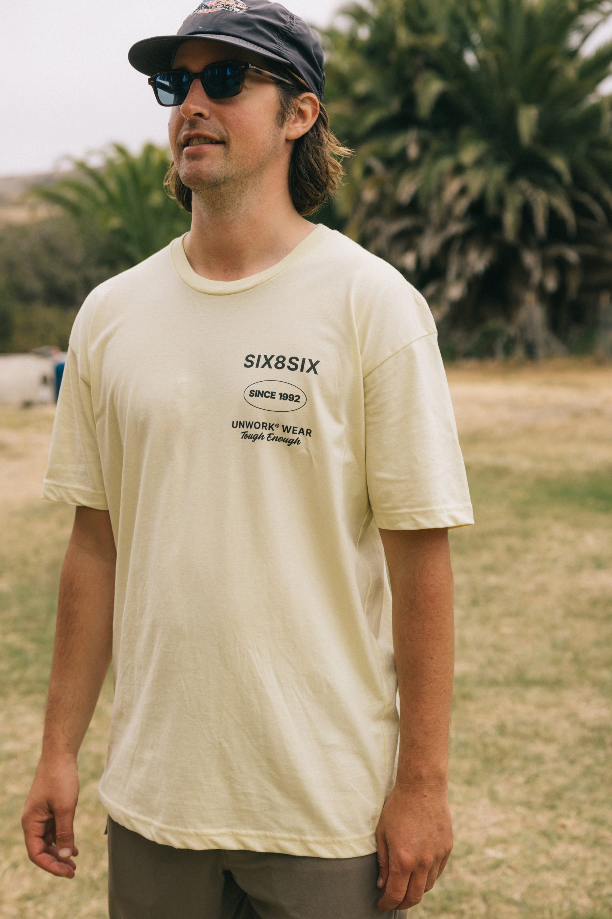 A man in sunglasses and a dark cap wears the 686 Unwork Wear Authentic S/S Tee by 686, standing outdoors on grass with palm trees and a blurred background.