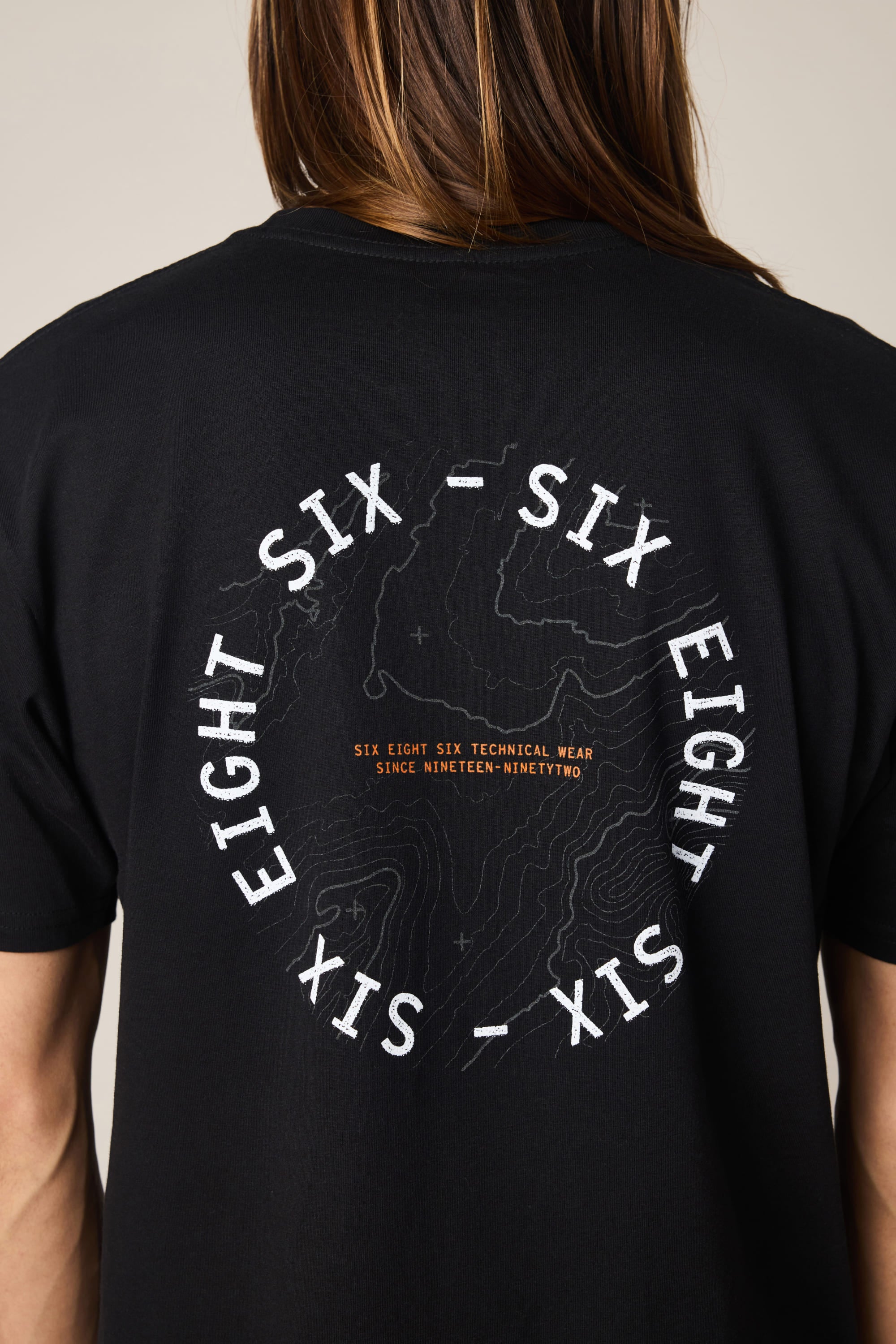 A person wears the 686 Lifts Authentic SS Tee, a men's cotton t-shirt by 686 featuring SIX EIGHT SIX in a circular contour map design and bold orange center text: SIX EIGHT SIX TECHNICAL WEAR SINCE NINETEEN-NINETY TWO.
