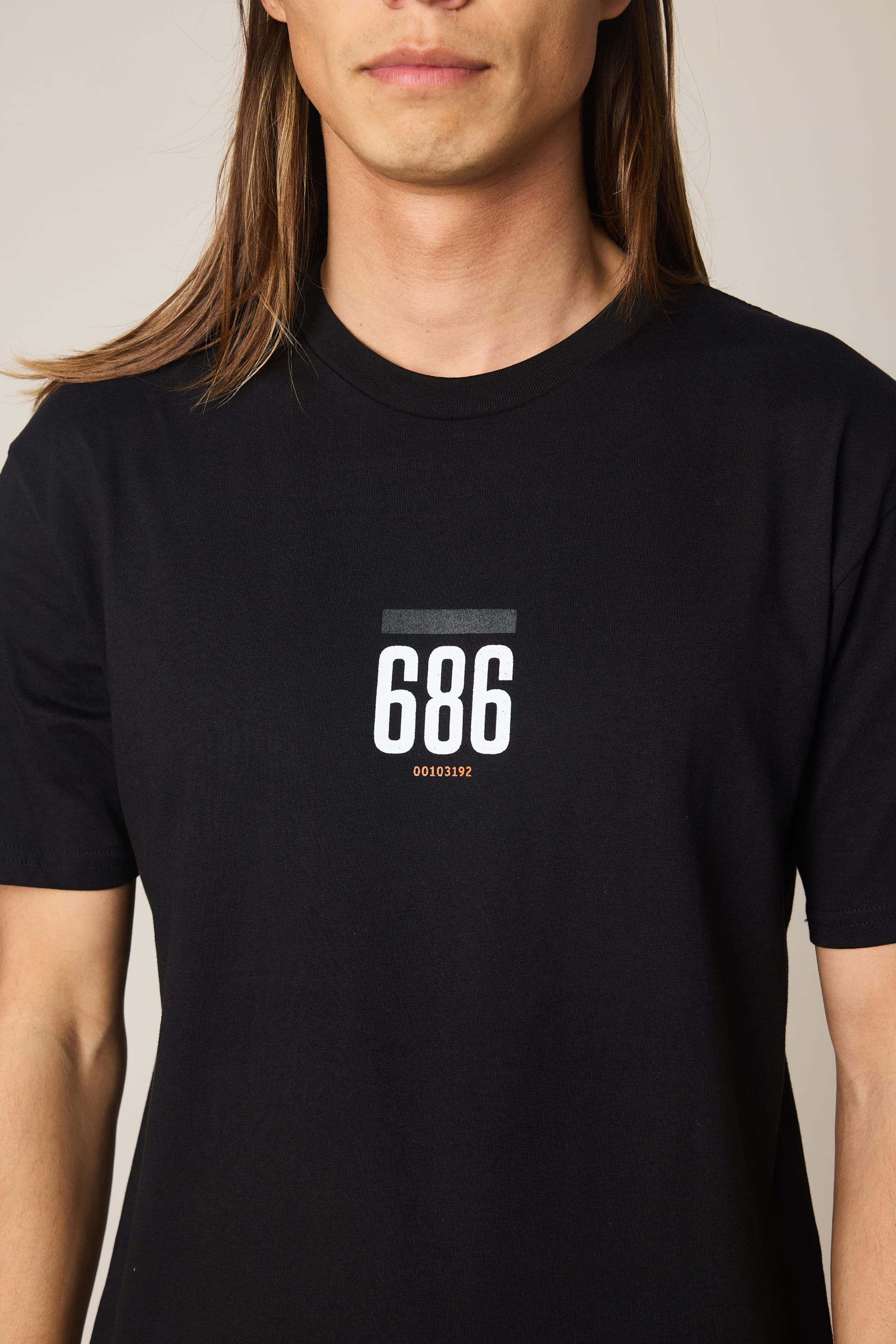 A person with long brown hair wears the 686 Lifts Authentic SS Tee by 686 in plain black, featuring "686" and code "0010192" printed in white and orange on the chest. The person's face is partially visible.