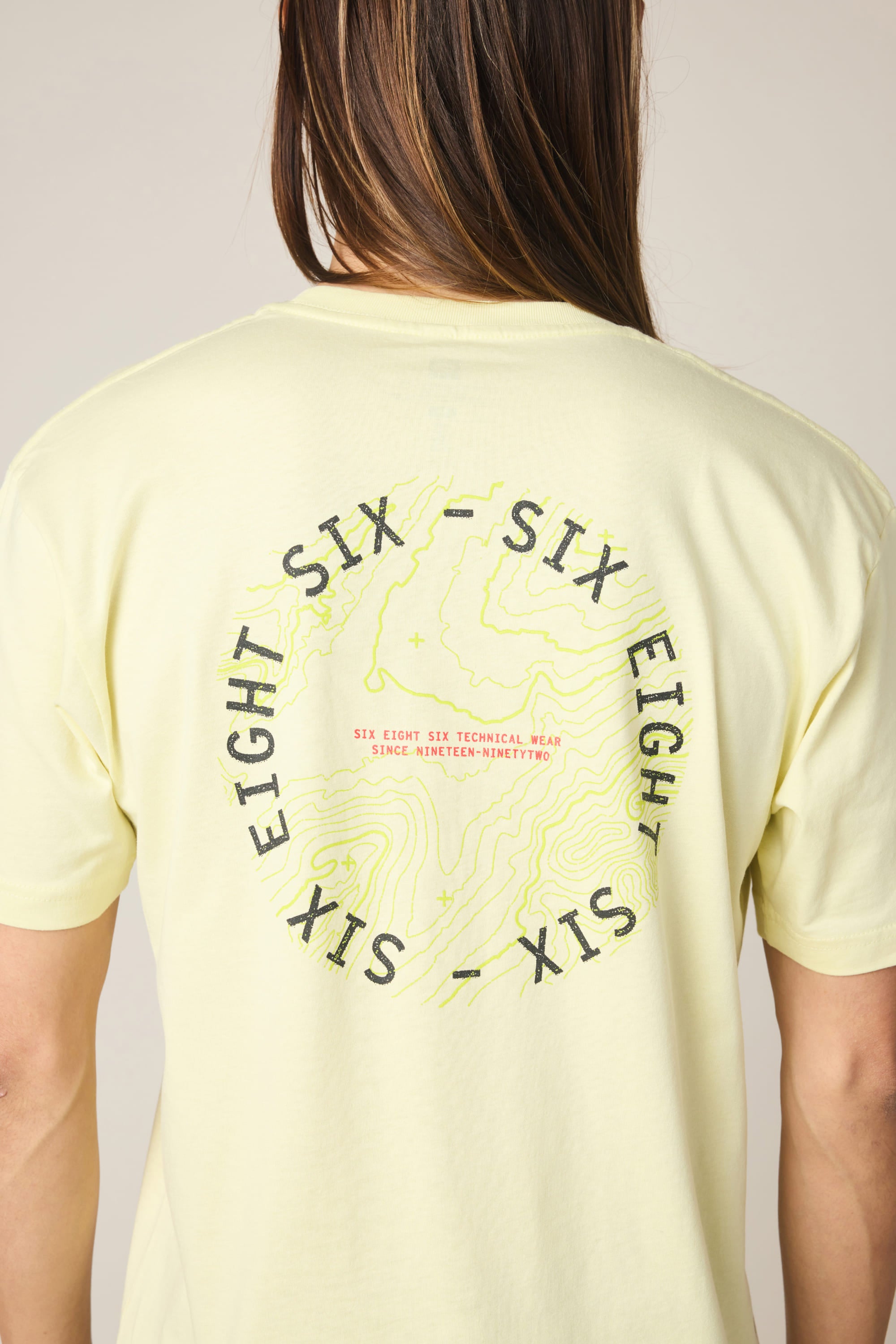 A person is shown from behind wearing the 686 Lifts Authentic SS Tee by 686, a light yellow 100% cotton men's t-shirt with a circular “SIX EIGHT SIX” and contour map graphic on the back, plus small red text in the center.