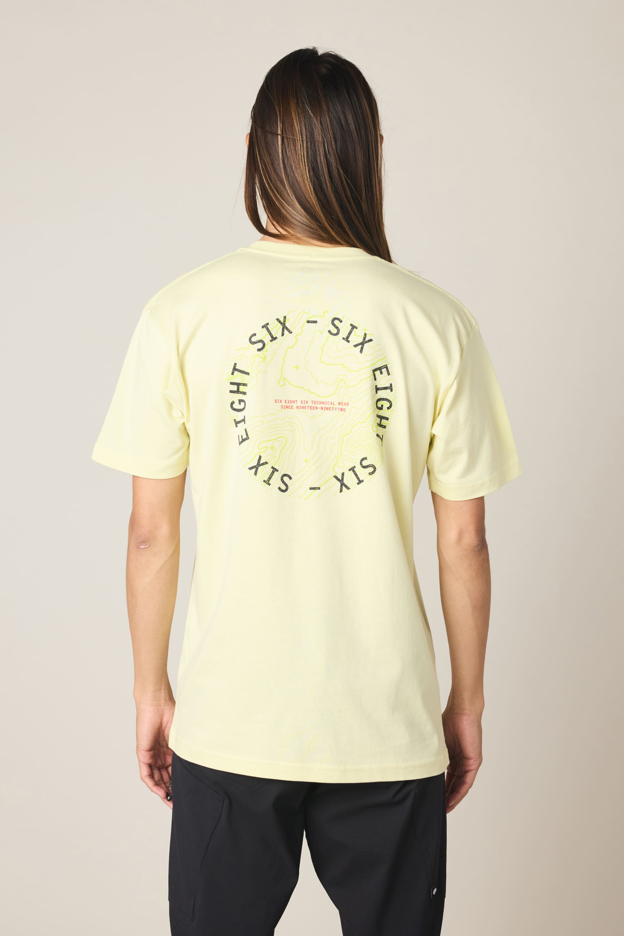 A person with long hair faces away from the camera, wearing the 686 Lifts Authentic SS Tee—a light yellow 100% cotton T-shirt by 686 featuring a circular “SIX EIGHT SIX” graphic and text on the back, set against a neutral background.