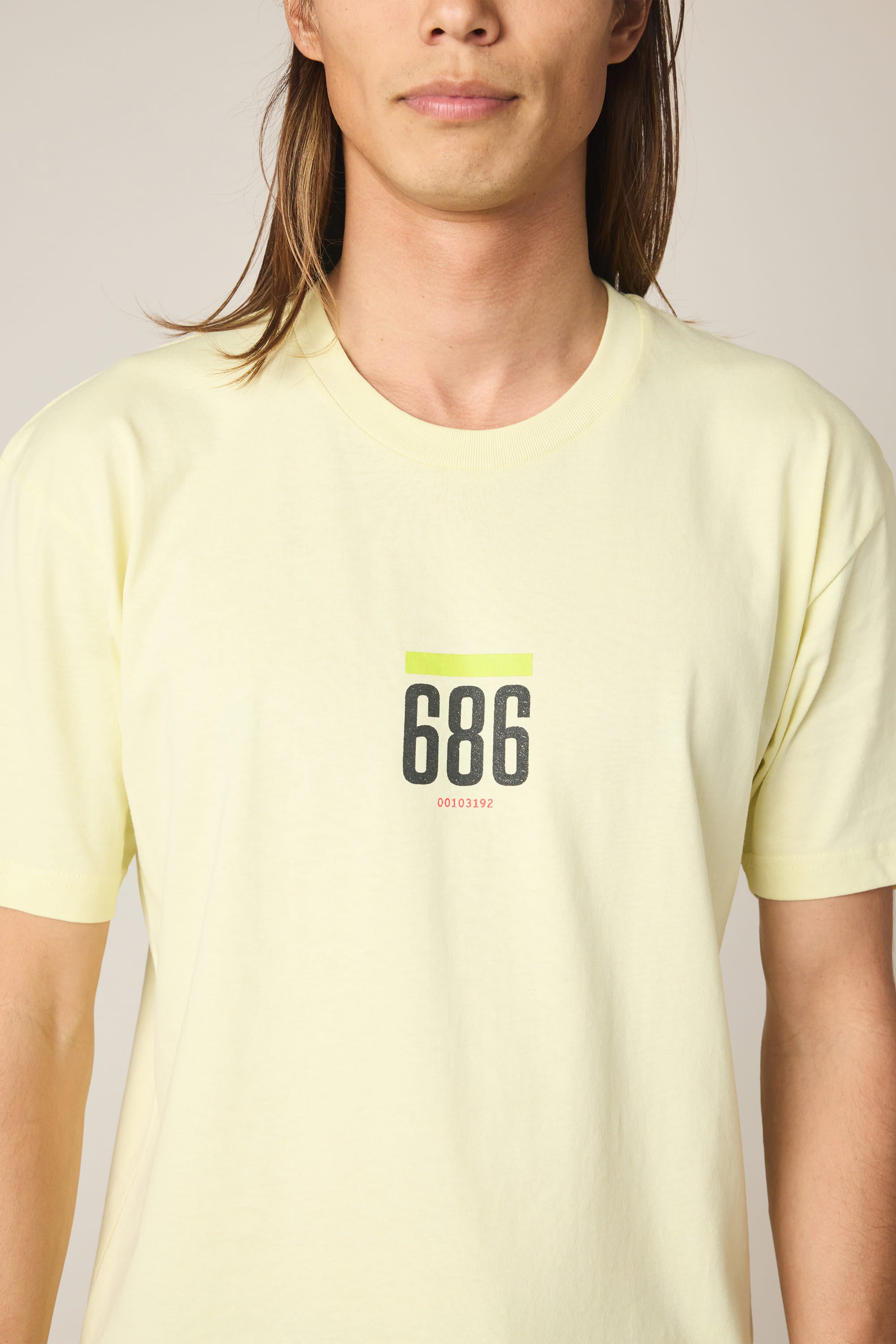 A person with long brown hair wears the 686 Lifts Authentic SS Tee by 686—a men’s light yellow cotton shirt with bold black “686,” a green line above, and small red numbers below, standing against a plain background.