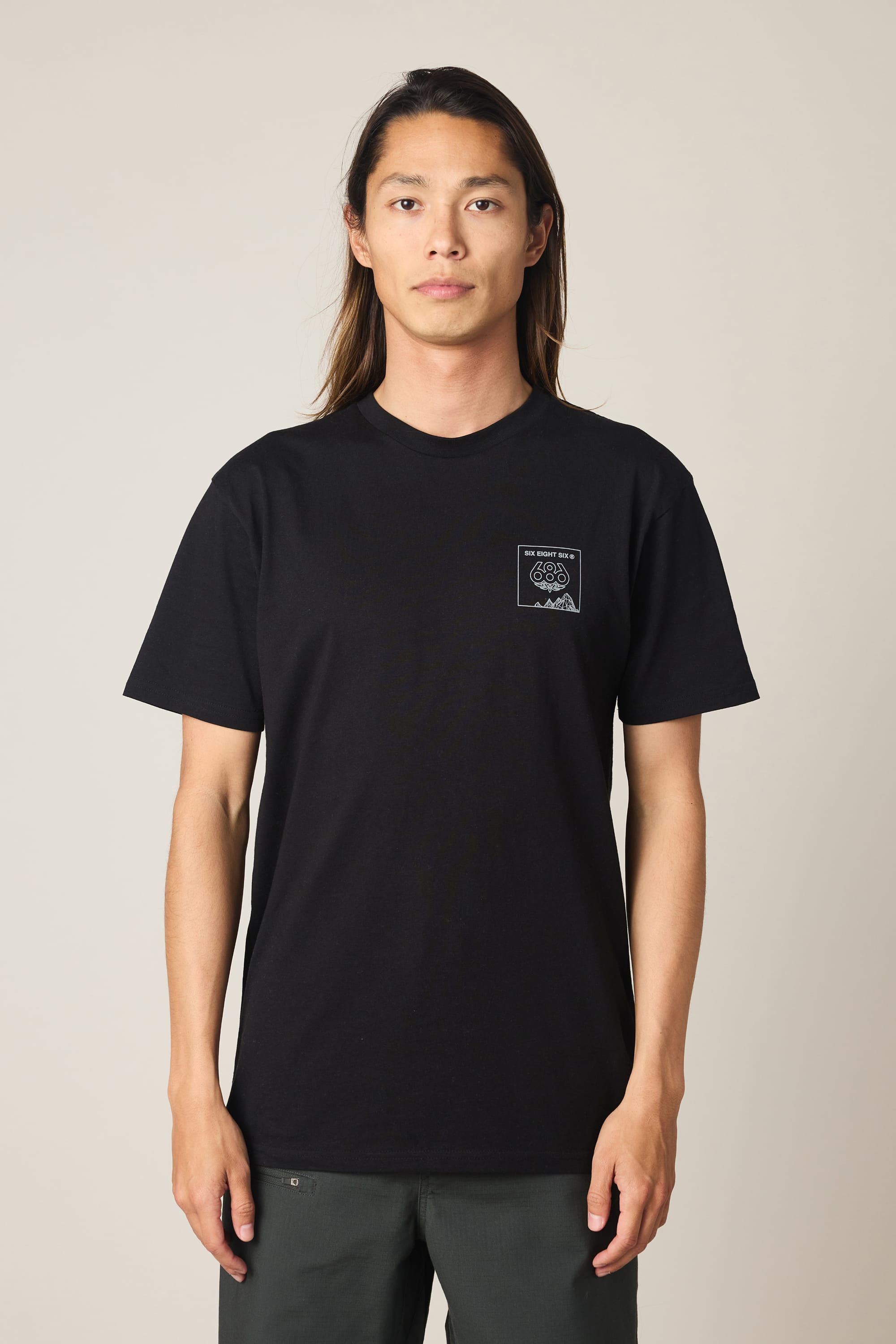 A person with long hair, wearing the 686 Cross-Cut Authentic S/S Tee by 686—a black 100% cotton men's t-shirt featuring a small white graphic on the left chest—and dark pants stands facing forward against a plain light background.