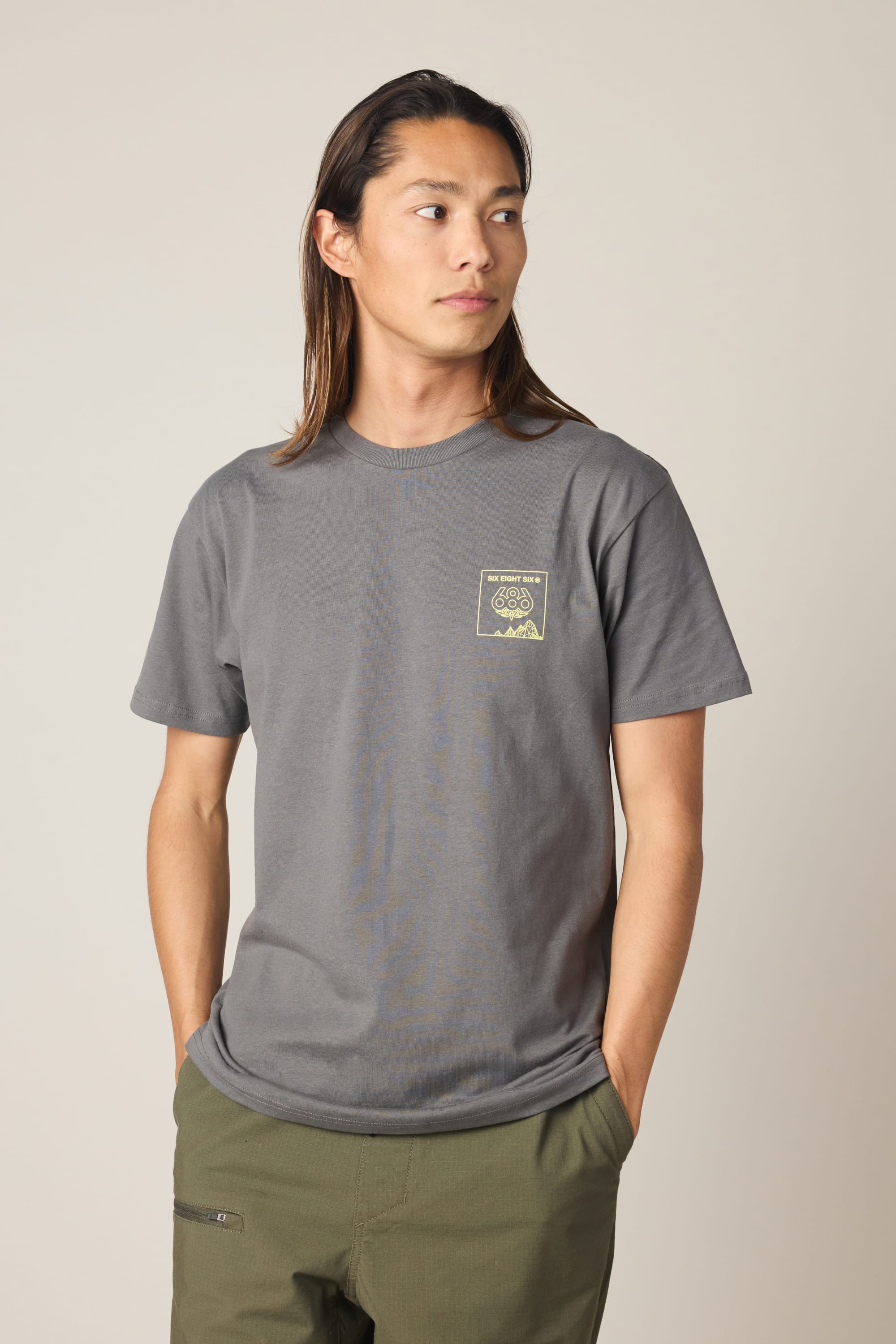 A person with long brown hair wears the 686 Cross-Cut Authentic S/S Tee in gray with a small yellow graphic on the left chest, paired with olive green pants, standing hands in pockets and looking to the side against a plain background.