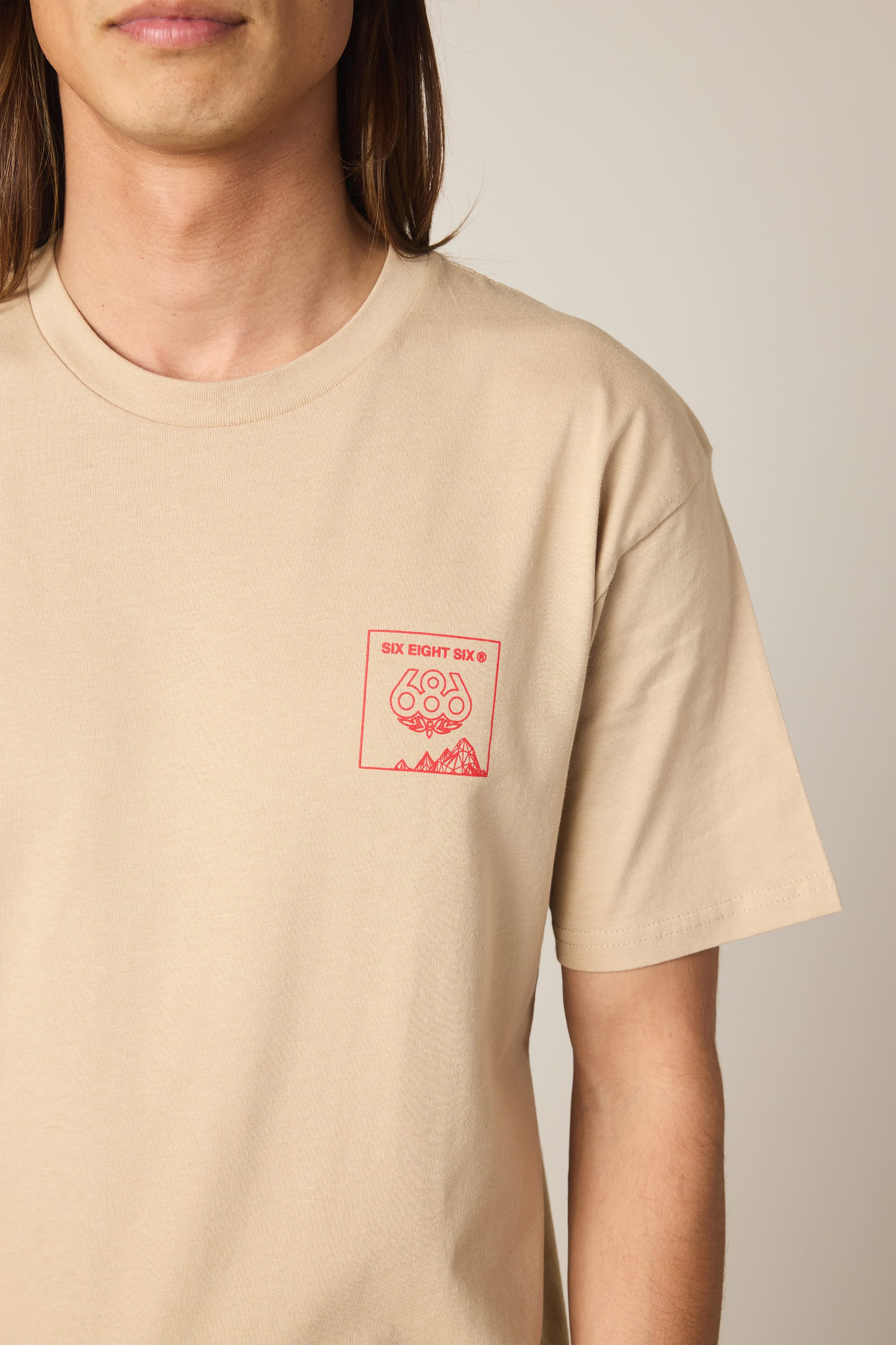 A person wearing the 686 Cross-Cut Authentic S/S Tee, a beige men's t-shirt by 686 featuring a small red graphic and the text "SIX EIGHT SIX" on the left chest. The 100% cotton shirt is shown from the upper torso with part of the person's face visible.