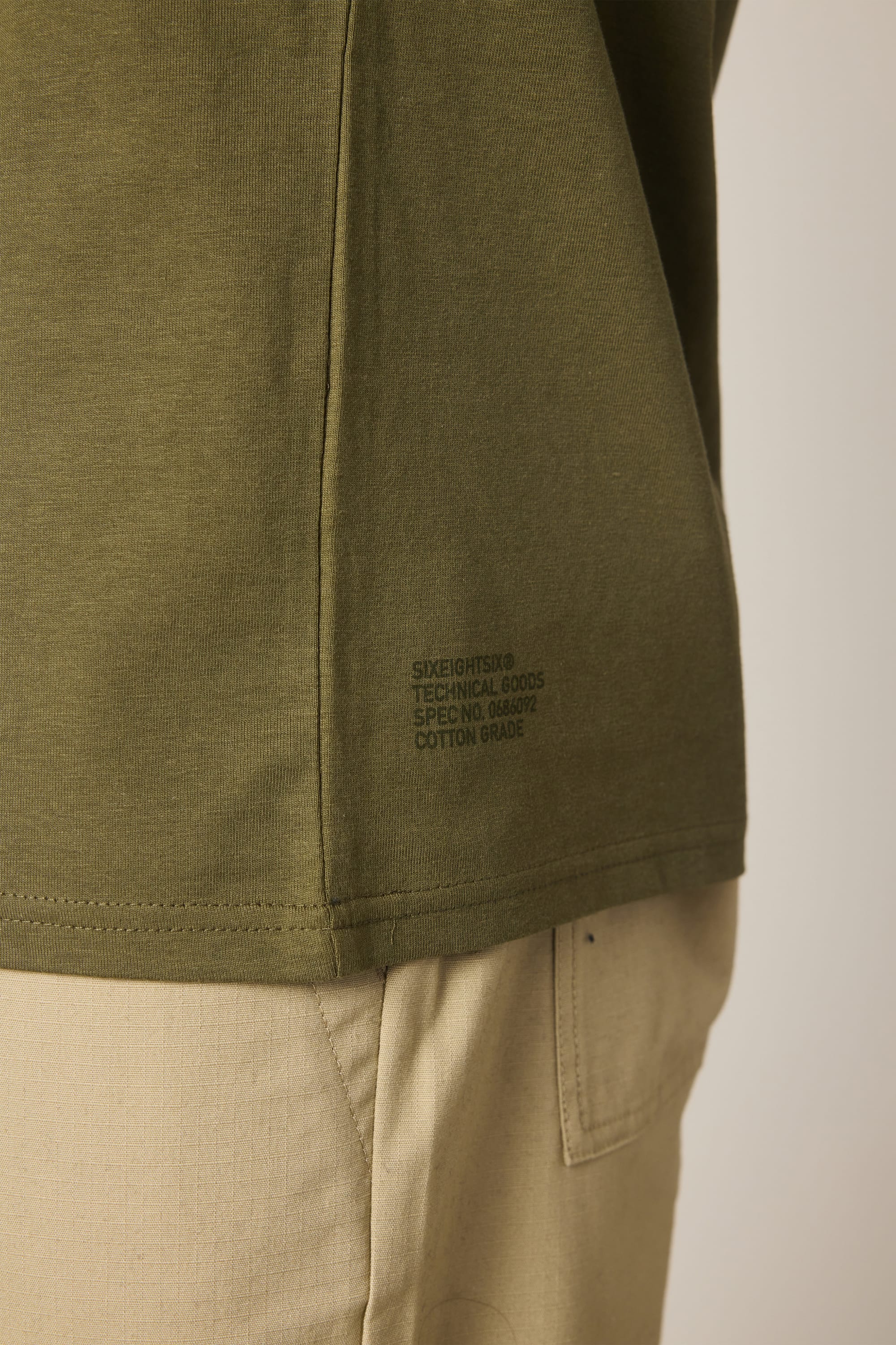 Close-up of a person wearing the 686 Knockout Authentic S/S Tee in olive green, featuring small printed text near the hem, paired with light beige pants, with the shirt’s side seam and the pocket of the pants visible.