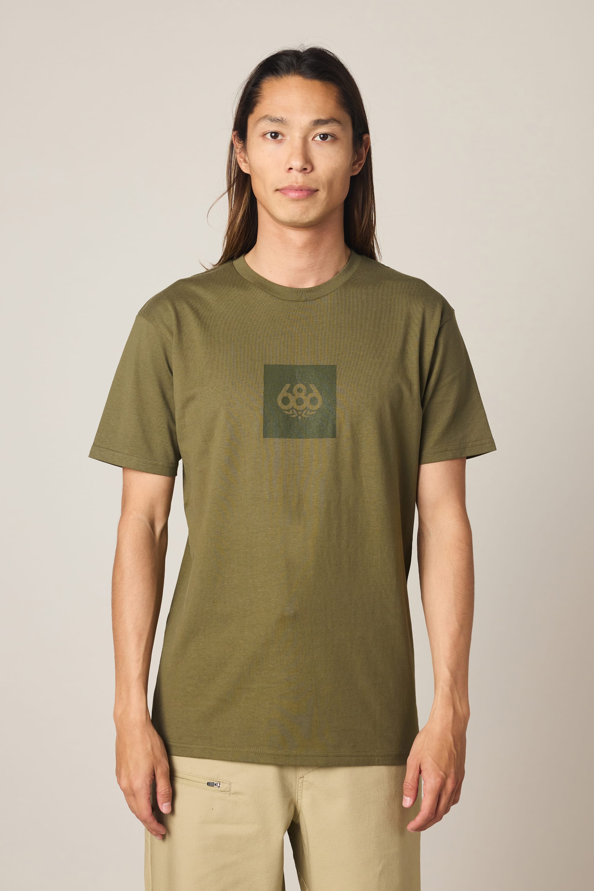 A person with long hair stands facing forward against a light background, wearing the 686 Knockout Authentic S/S Tee in olive green—featuring a subtle dark green square logo—and beige pants.