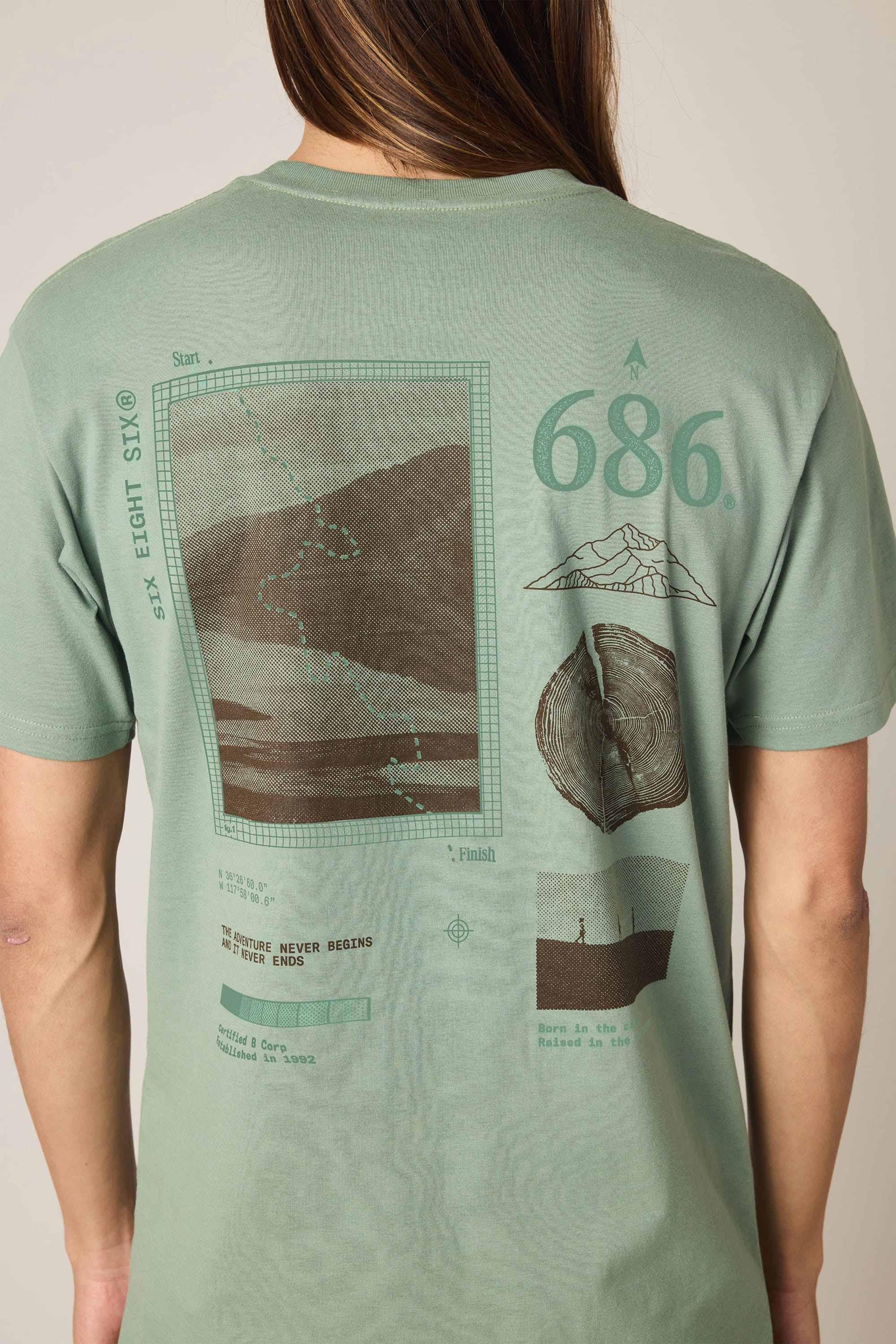 Wearing the 686 Path Authentic S/S Tee, a sage green 100% cotton t-shirt by 686, featuring graphics of a mountain, tree ring, landscape, dotted path, the number 686, and the quote "The journey never begins and never ends.