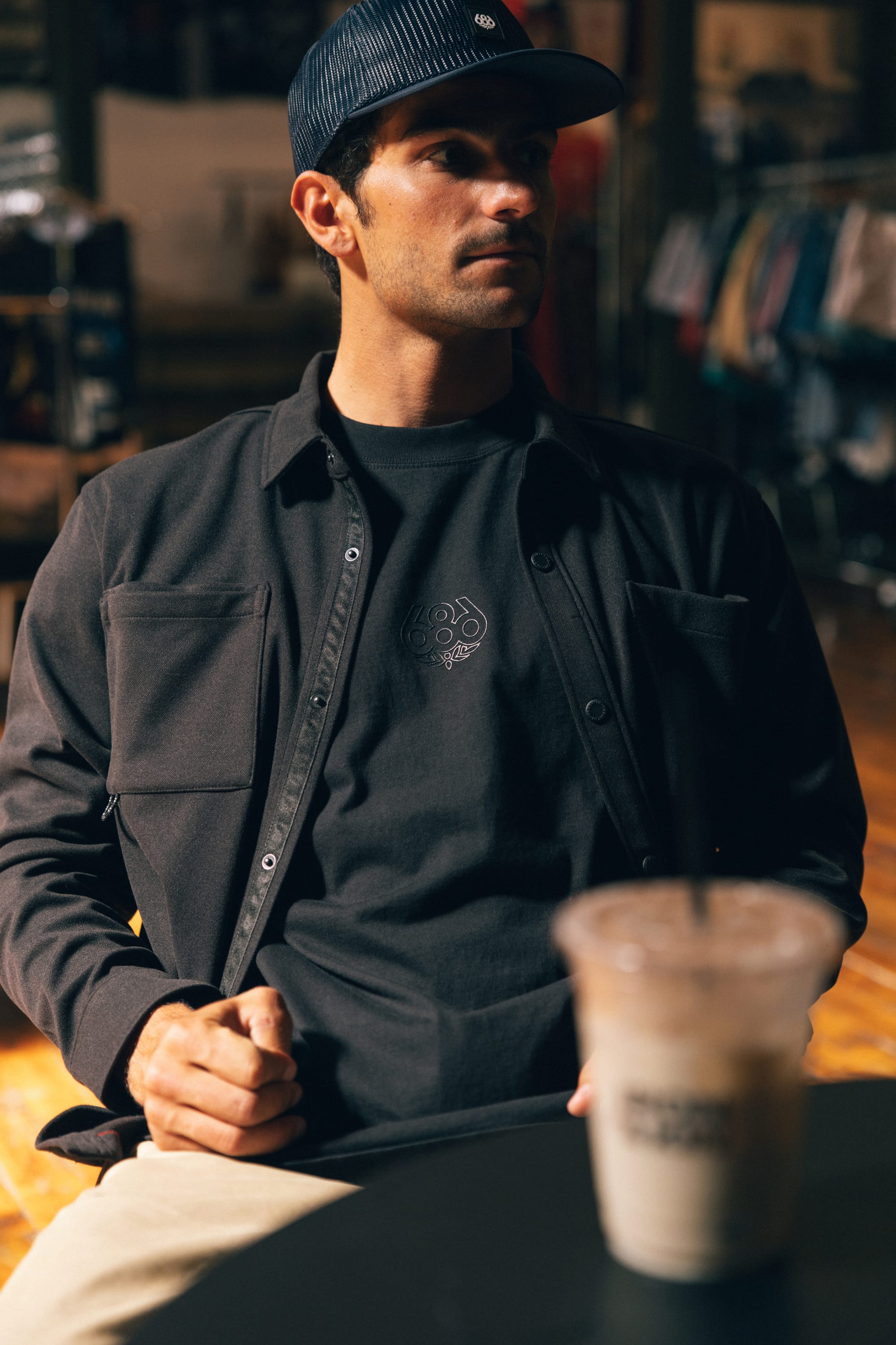 A man in a dark jacket and cap sits indoors at a table with an iced coffee in the foreground, his comfortable 686 High Def Relaxed S/S Tee from 686—crafted from 100% cotton—peeking out beneath his jacket. Blurred clothing racks are visible in the background.