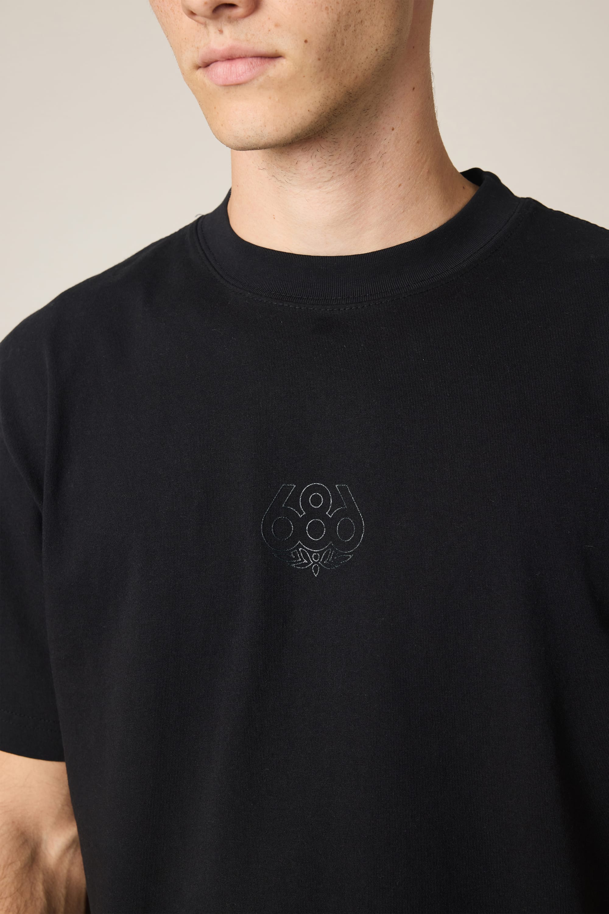 A person wears the 686 High Def Relaxed S/S Tee in black, featuring a small, subtle 686 logo on the chest. The photo is cropped to show only the lower half of the face and upper torso against a neutral background. This classic cotton t-shirt from 686 provides both comfort and understated style.