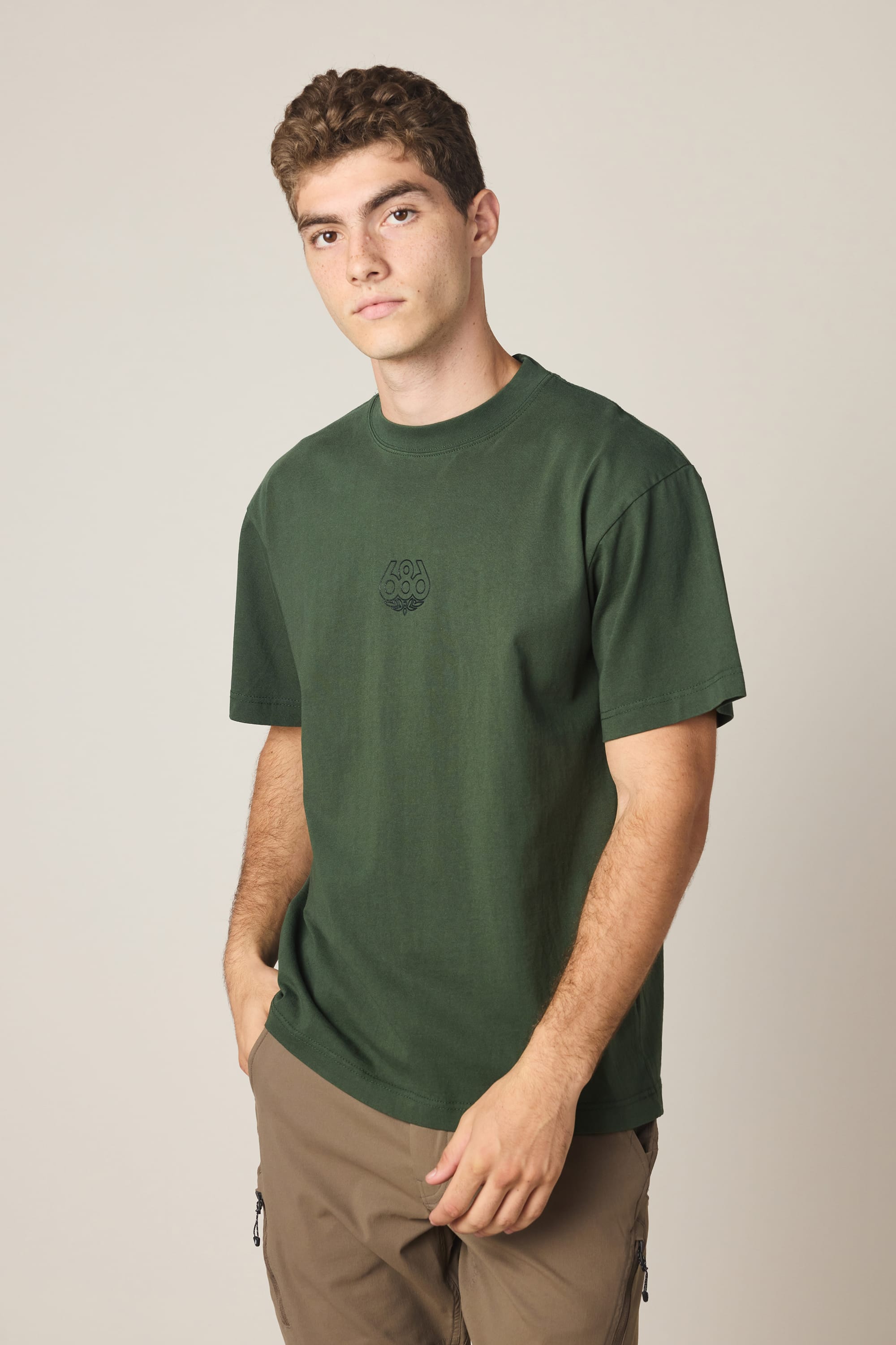 A young man with short curly hair wears the 686 High Def Relaxed S/S Tee by 686 in dark green and brown pants, standing against a plain light-colored background with one hand in his pocket.