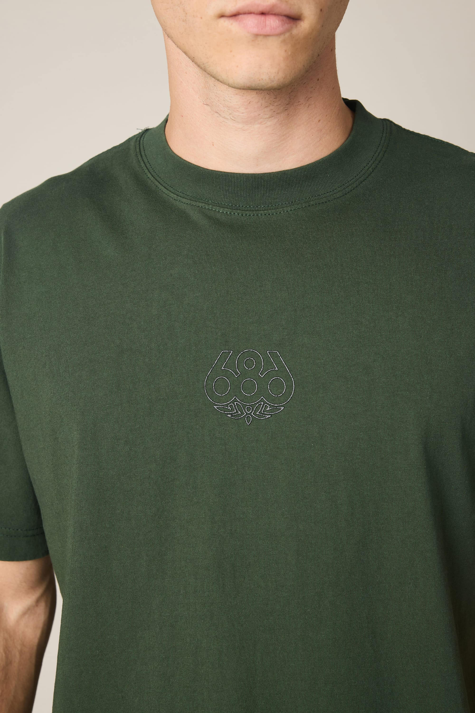 A person wearing the 686 High Def Relaxed S/S Tee by 686—a dark green men's t-shirt made from 100% cotton and featuring a small, subtle embroidered 686 logo with a leaf design on the chest. The person's face is partially out of frame.