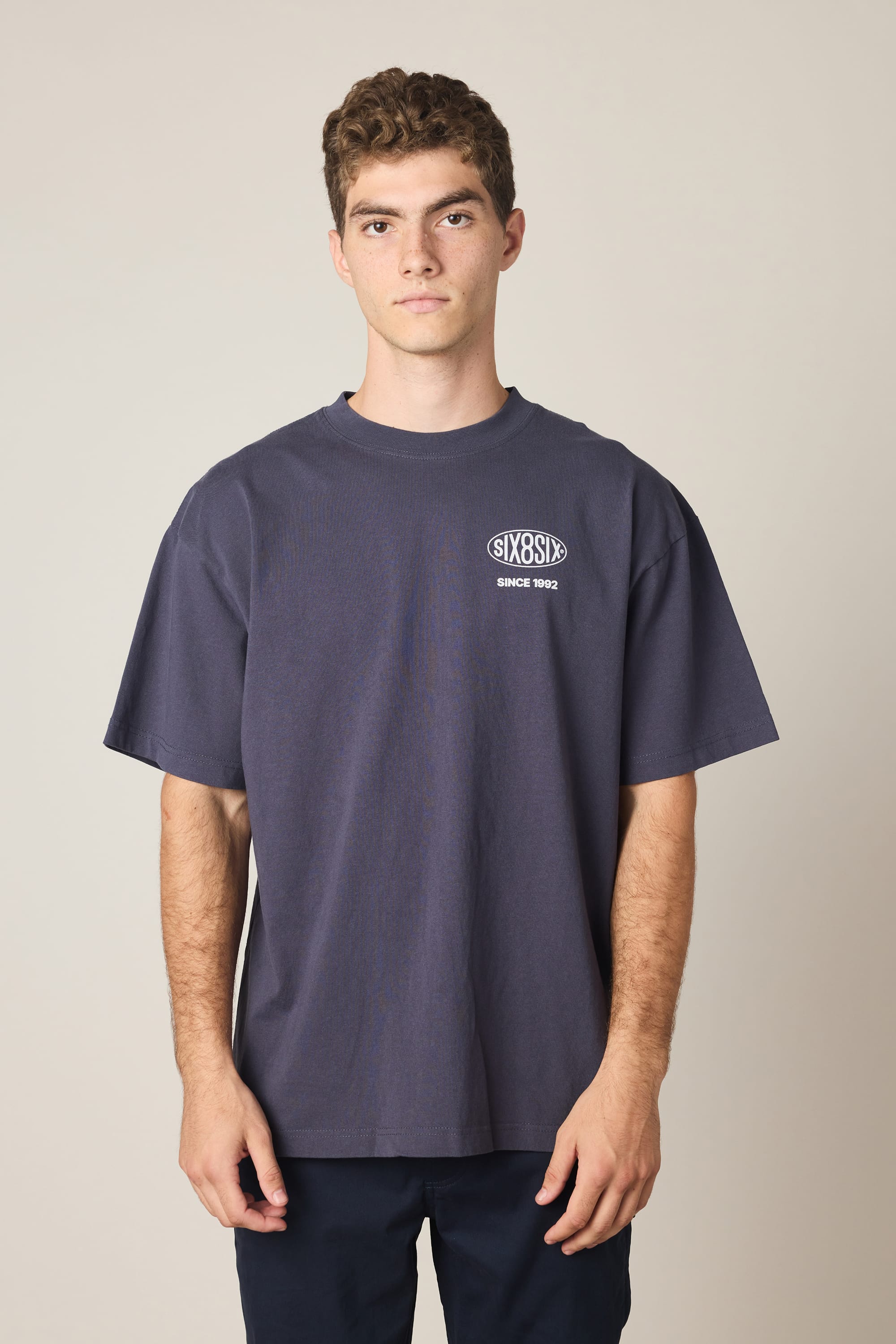 A young man with short curly hair stands against a beige background wearing the 686 92 Relaxed S/S Tee by 686, a loose-fitting men's t-shirt made from 100% cotton in dark gray, featuring "SIXSIX" and "SINCE 1993" printed on the upper left chest.