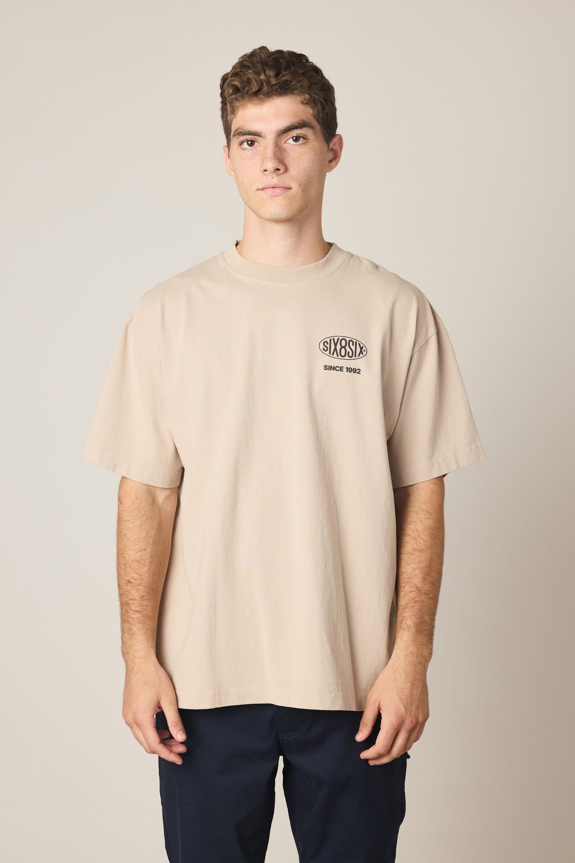 A young man with short curly hair is seen standing against a plain light background, wearing the 686 92 Relaxed S/S Tee by 686—an oversized men’s beige t-shirt featuring "SIXSIX" and "SINCE 1993" printed on the chest. This stylish tee is crafted from 100% cotton for all-day comfort.