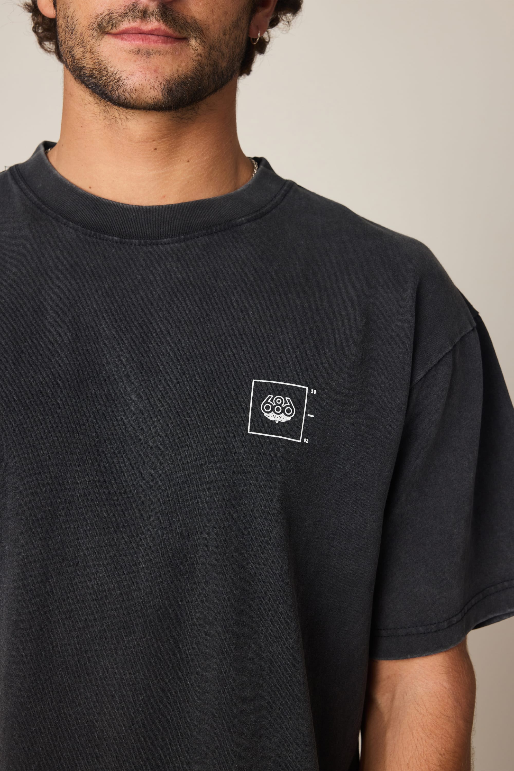 A man wears the 686 Peaks Relaxed SS Tee, a faded black men's t-shirt by 686 featuring a small white geometric logo in a square on the left chest. Only his lower face and upper torso are visible. Made from 100% cotton for comfort.