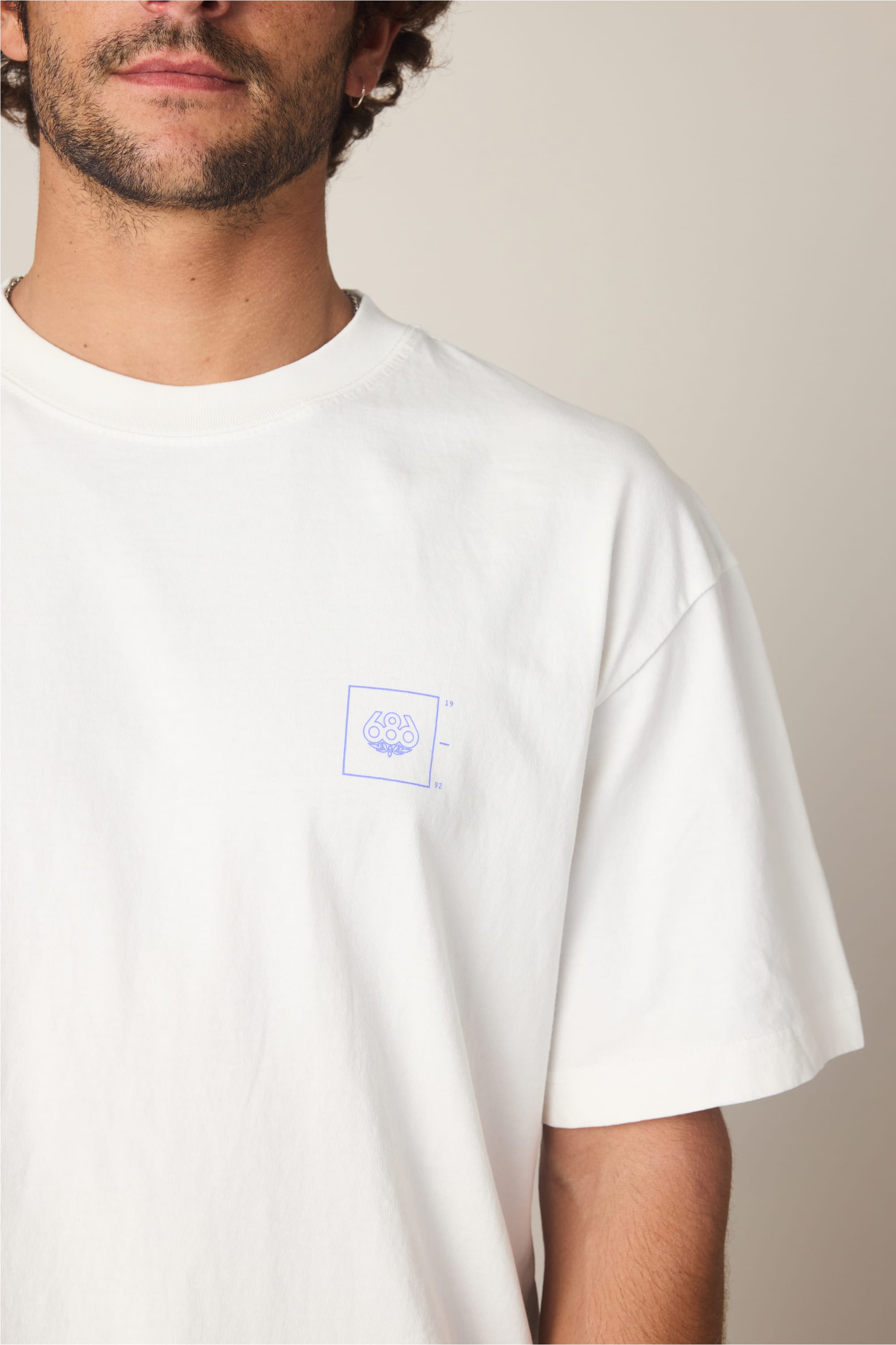 A person wears the 686 Peaks Relaxed SS Tee by 686—a plain white men’s t-shirt made from 100% cotton with a small blue graphic logo on the left chest. Only their lower face and upper torso are visible.