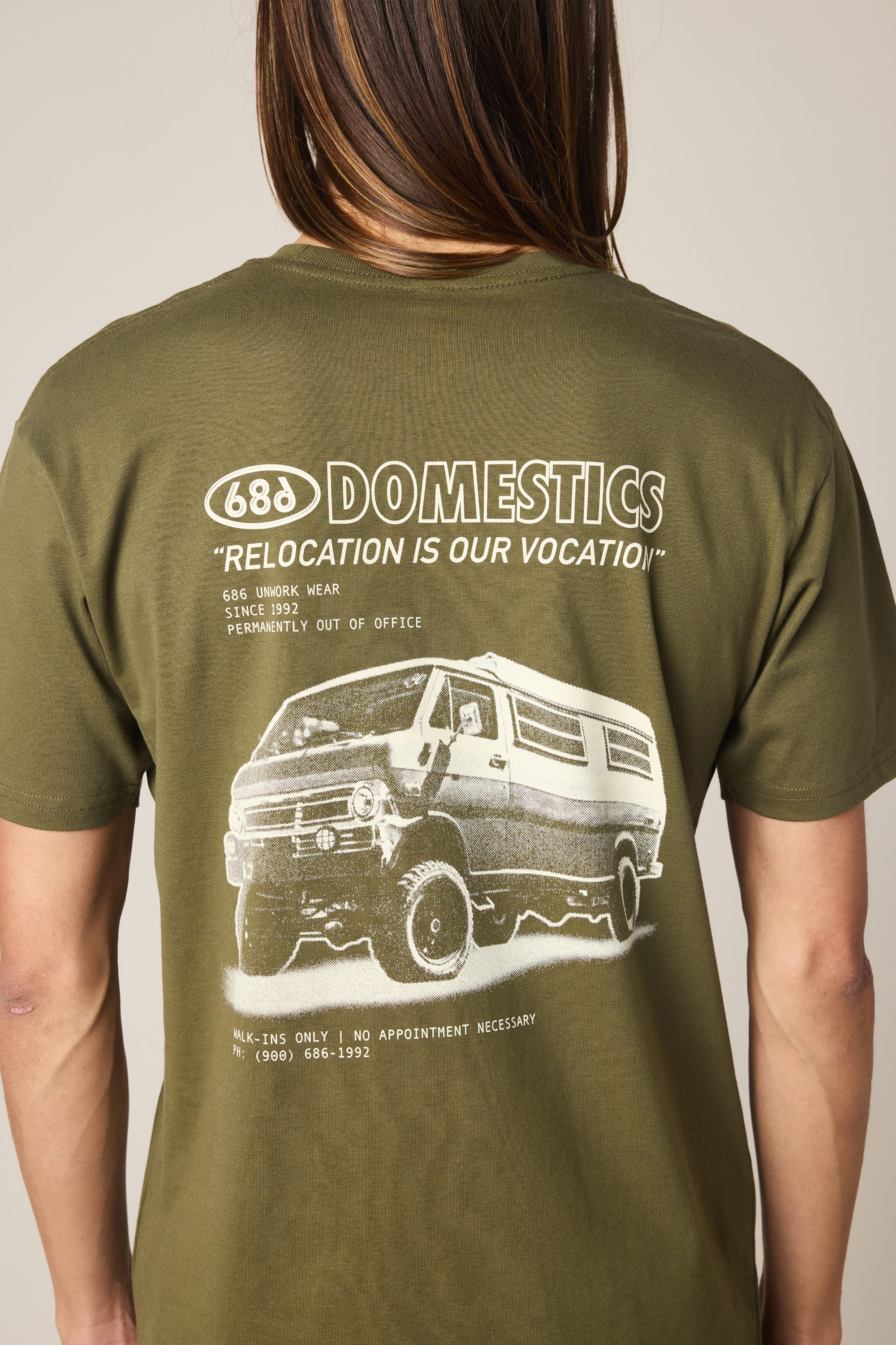A person wears the 686 Men's Domestics Authentic SS Tee, an olive green 100% cotton shirt featuring a van graphic with “DOMESTICS ‘RELOCATION IS OUR VOCATION’” and smaller white text on the back for all-day comfort.