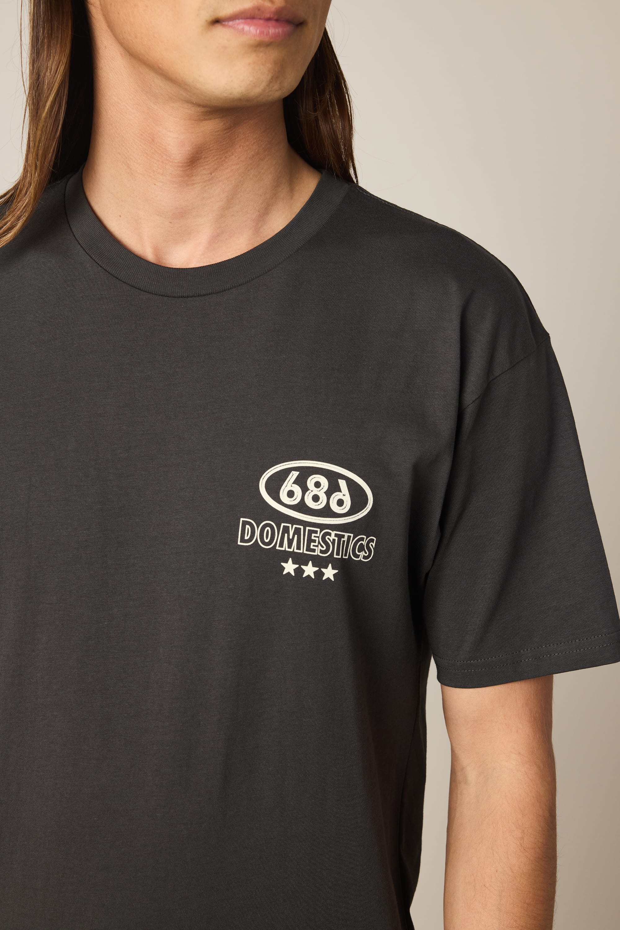 A person is wearing the 686 Men's Domestics Authentic SS Tee—a black, classic-fit t-shirt made from 100% cotton by 686. The design features "68d DOMESTICS" and three white stars printed on the upper left chest. The image captures the shirt from shoulders to just above the elbow, emphasizing its comfortable fit.