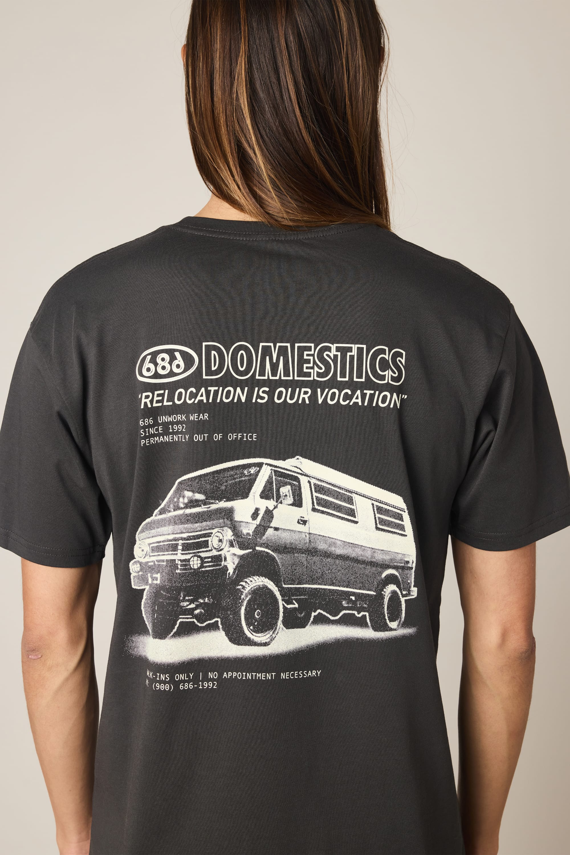 A person wearing the 686 Men's Domestics Authentic SS Tee—a black 100% cotton T-shirt by 686 with white graphics on the back, featuring a van, “688 DOMESTICS RELOCATION IS OUR VOCATION,” and detailed text.