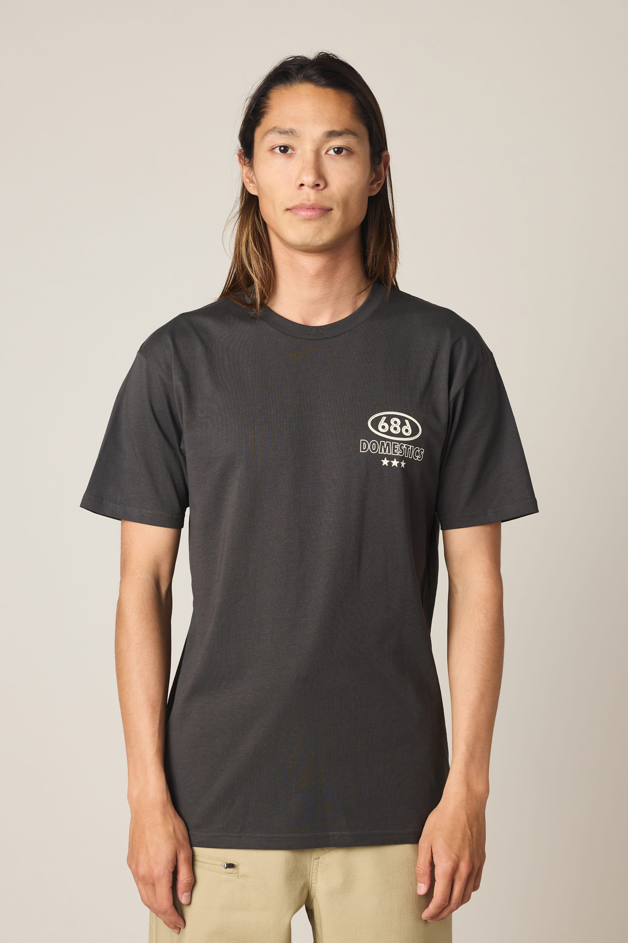 A person with long brown hair faces the camera, wearing the 686 Men's Domestics Authentic SS Tee—a dark gray 100% cotton t-shirt by 686 with a small white Domestics logo and two stars on the left chest. The background is plain and light.