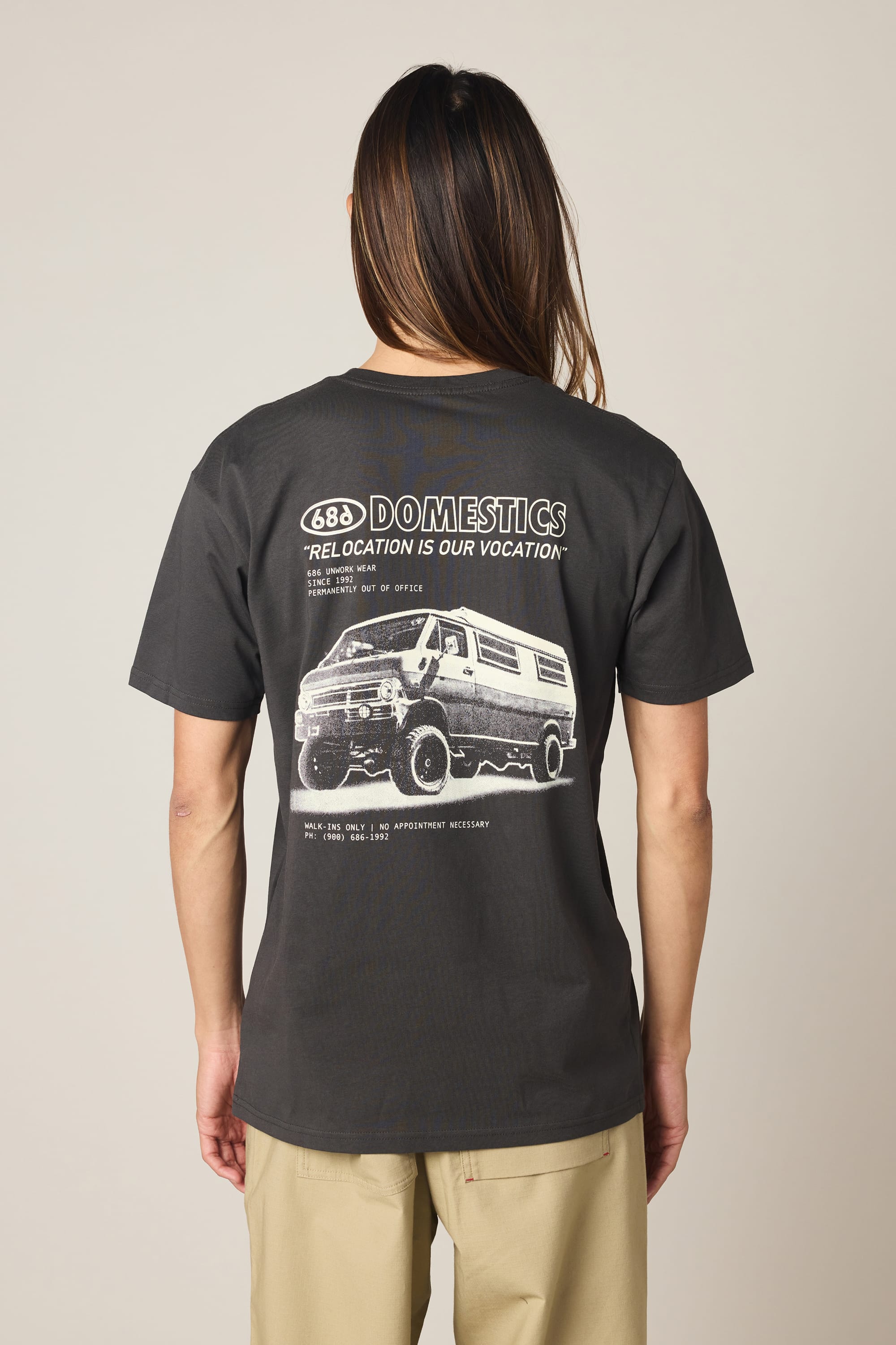 A person is shown from behind wearing the 686 Men's Domestics Authentic SS Tee by 686—a black, 100% cotton men’s t-shirt featuring a van graphic and the phrase “688 DOMESTICS ‘RELOCATION IS OUR VOCATION’”—paired with beige pants and set against a plain light background.